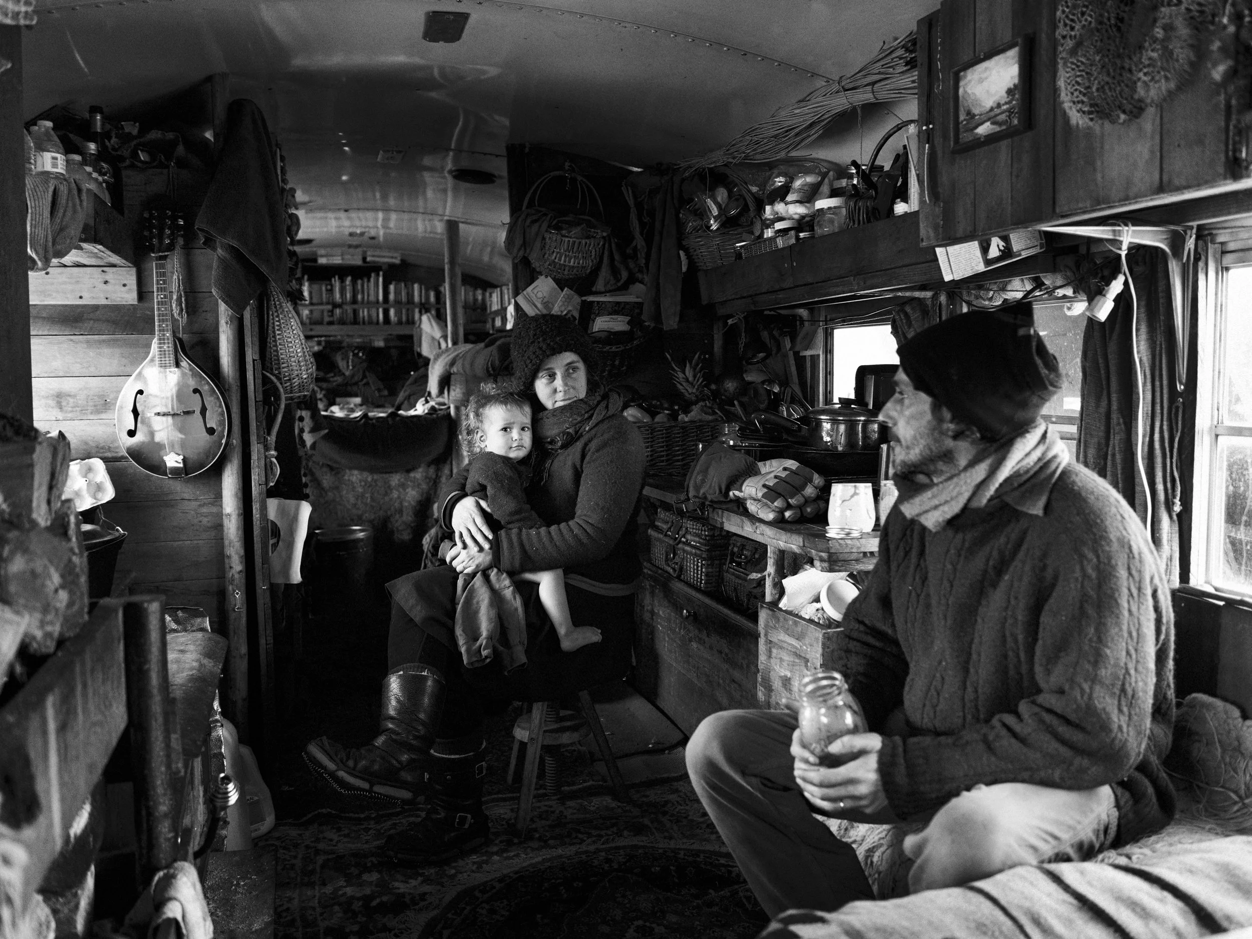 Black and white documentary photograph of Ande and Kayla Blanchflower sitting in the cluttered, narrow living space inside a converted school bus at the Standing Rock camp, with clothes, bags, and winter supplies hanging from the ceiling and walls.
