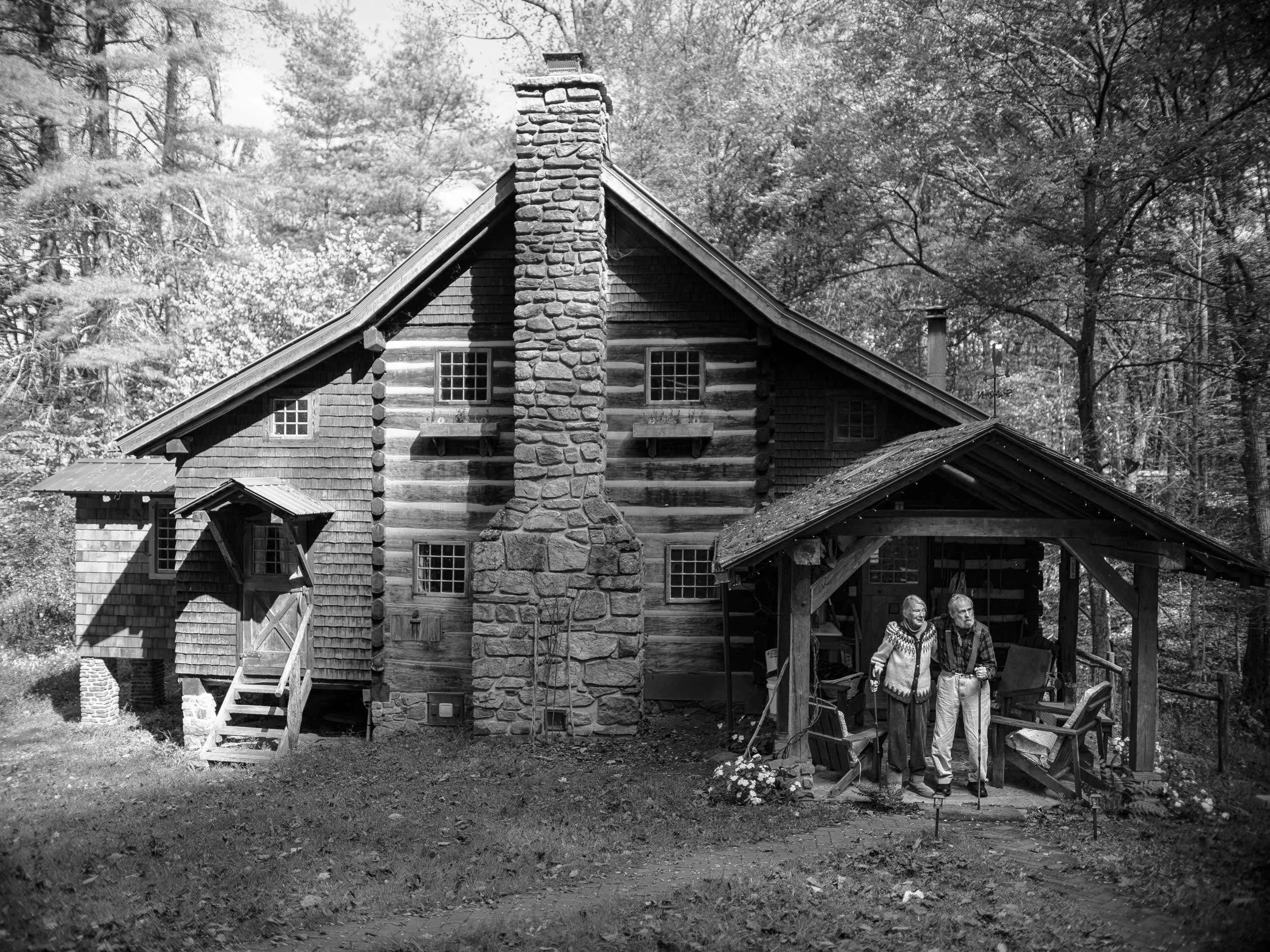 A wide black and white documentary photograph of a large two-story rustic log cabin with a central stone chimney, featuring an elderly couple standing together under the front porch awning in a wooded Western North Carolina setting.