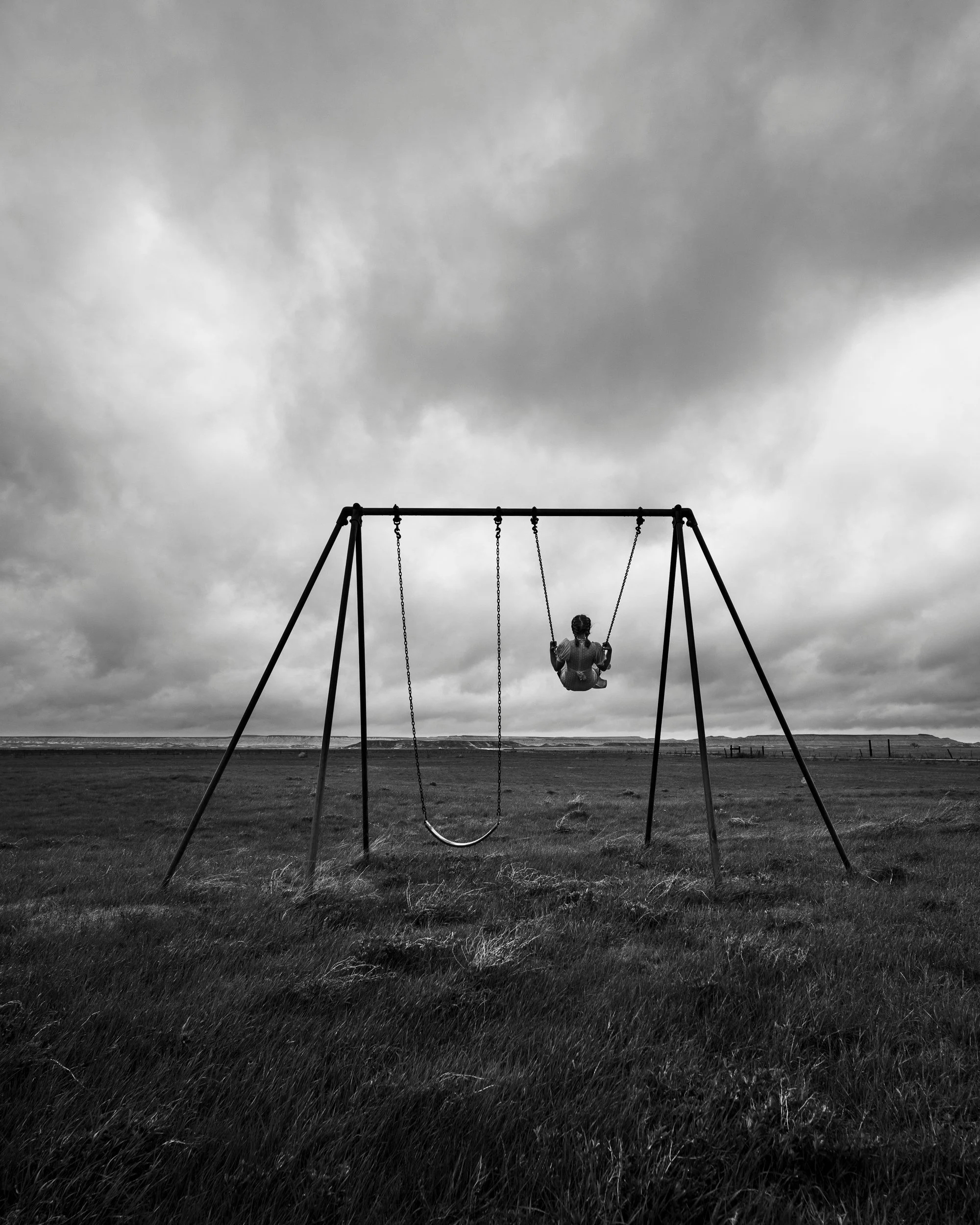 A dramatic black and white fine art photograph of a lone person high in mid-swing on a minimalist metal swing set, captured against a vast, empty field and a massive, heavy blanket of dark storm clouds in South Dakota.