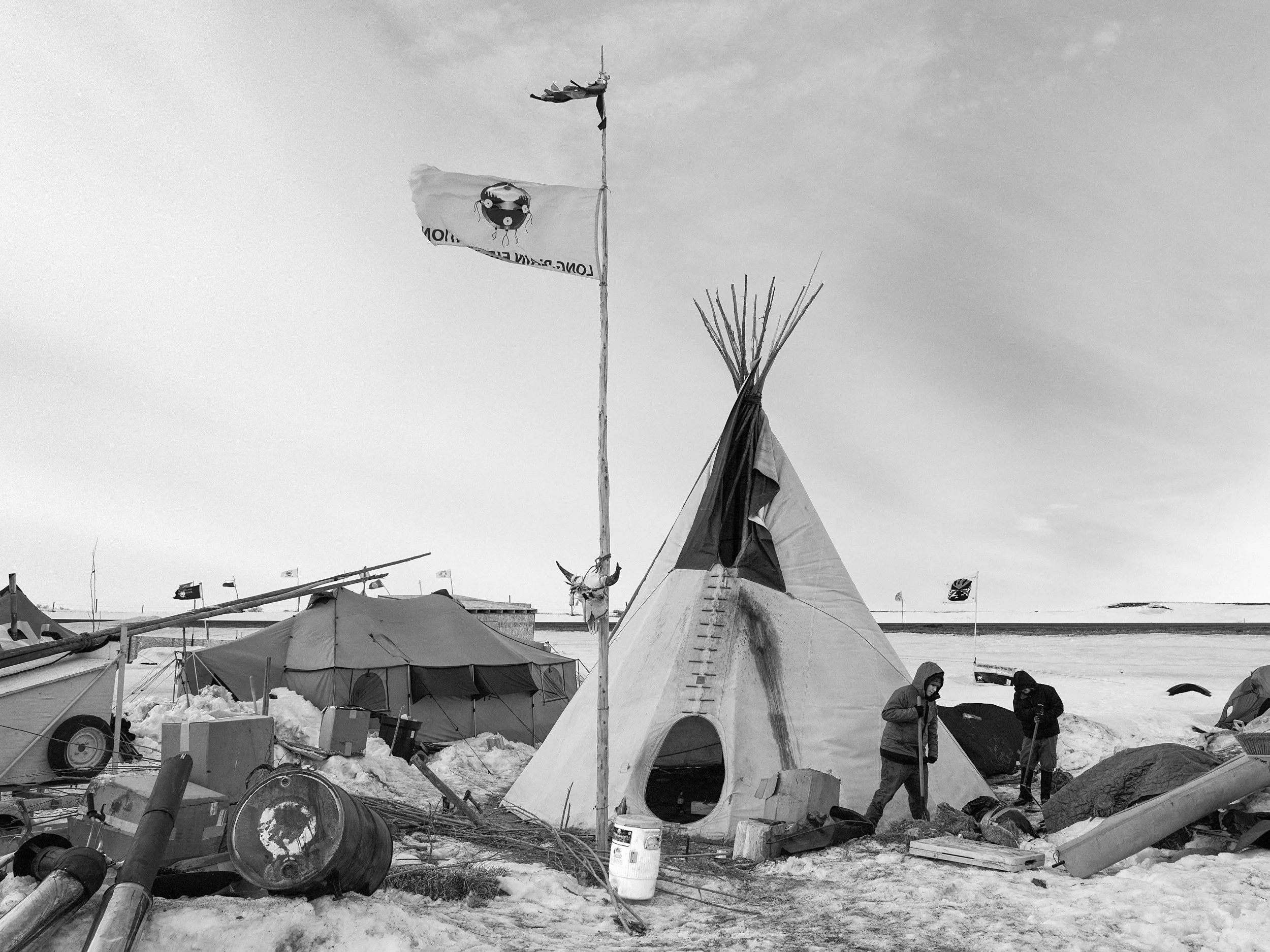 Black and white documentary photograph looking up at a white flag featuring a graphic emblem flying from a tall flagpole, with the bare wooden support poles of a traditional tipi rising into the sky just behind it.