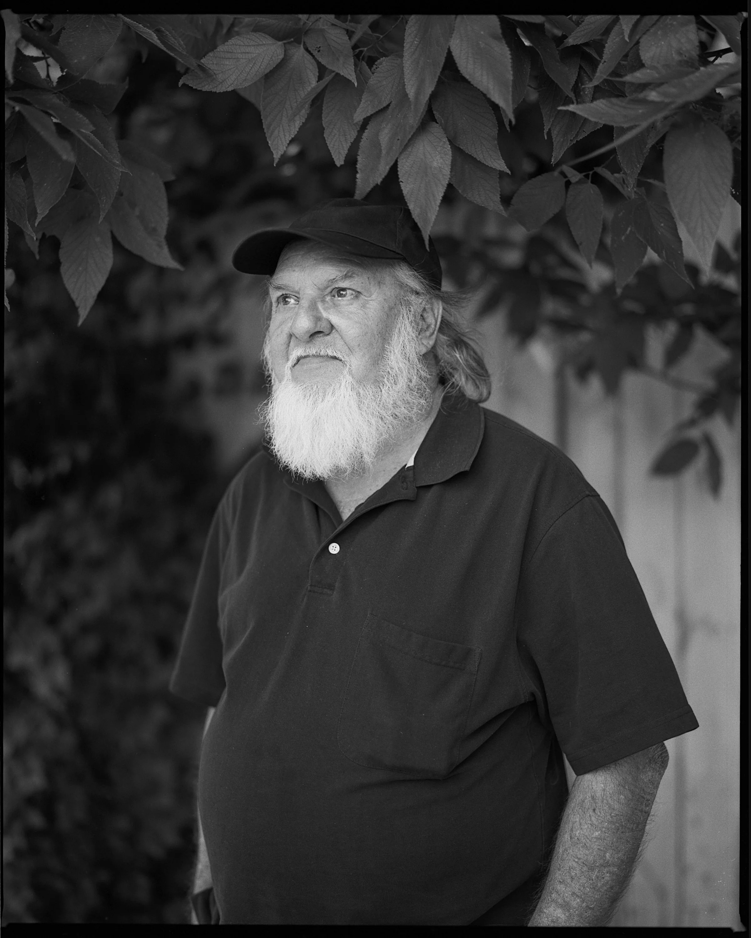 A timeless black and white environmental portrait of an older man with a long white beard and a baseball cap standing under lush tree leaves, from the One Way Out, Lord documentary series by Parker J Pfister.