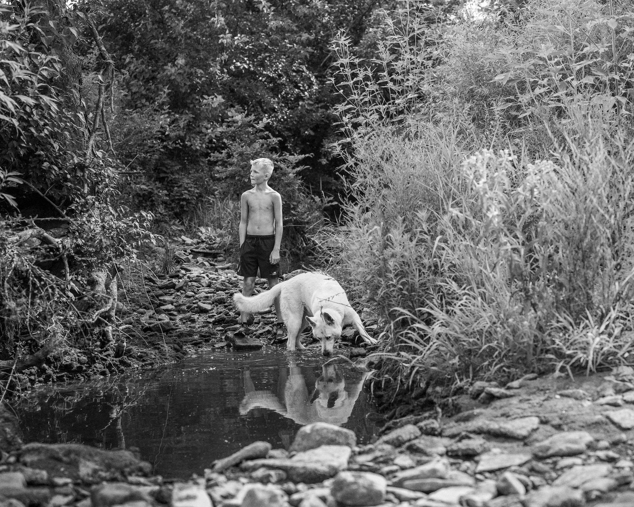 A beautiful black and white environmental photograph of a young boy standing in a rocky creek beside a white dog drinking from the water, surrounded by dense summer foliage, from One Way Out, Lord by Parker J Pfister.
