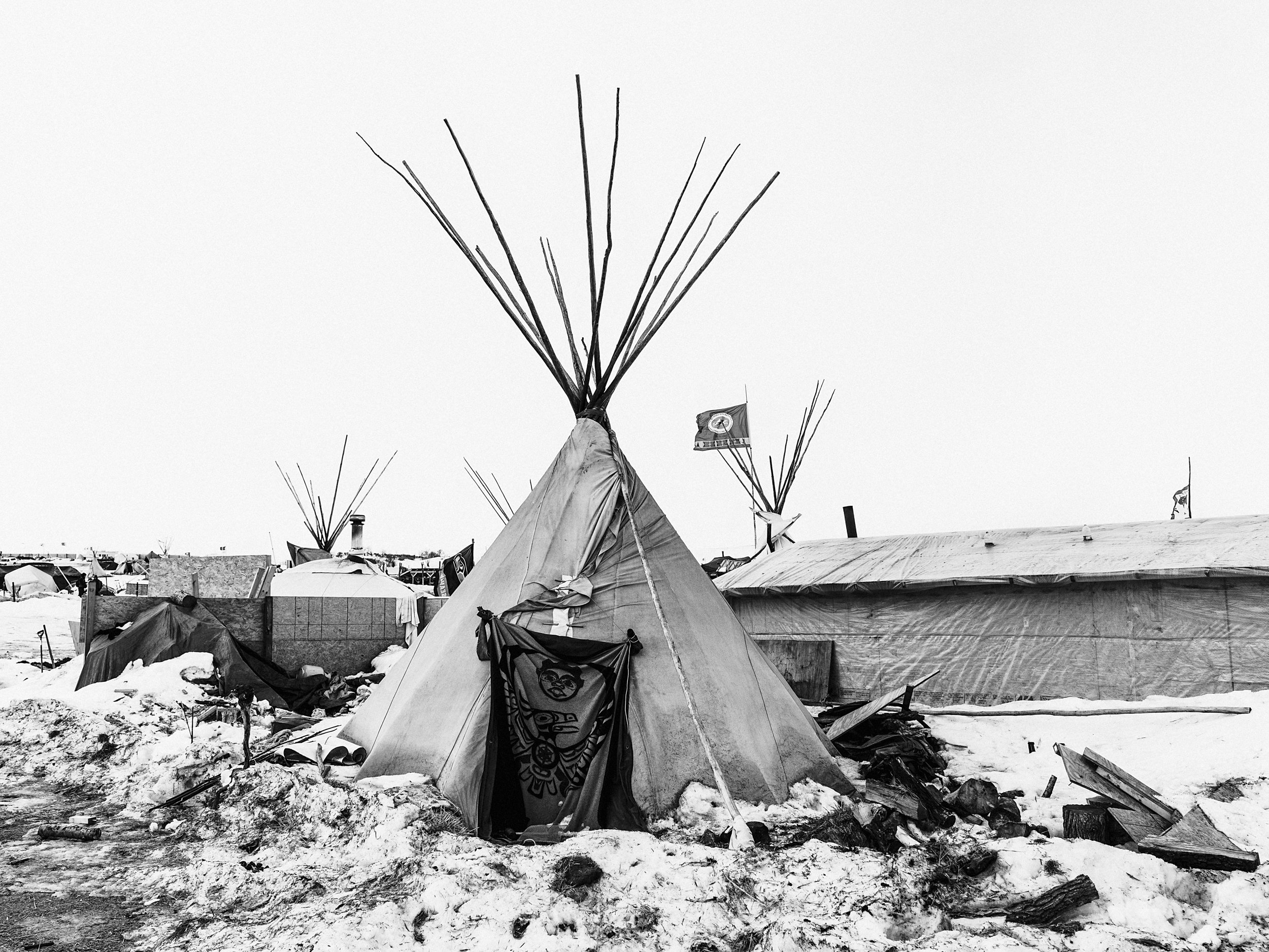 Black and white documentary photograph of a large pile of discarded tarps, gear, and debris sitting on a patch of snowy, muddy ground at the Standing Rock camp, with a large dump truck parked in the background.