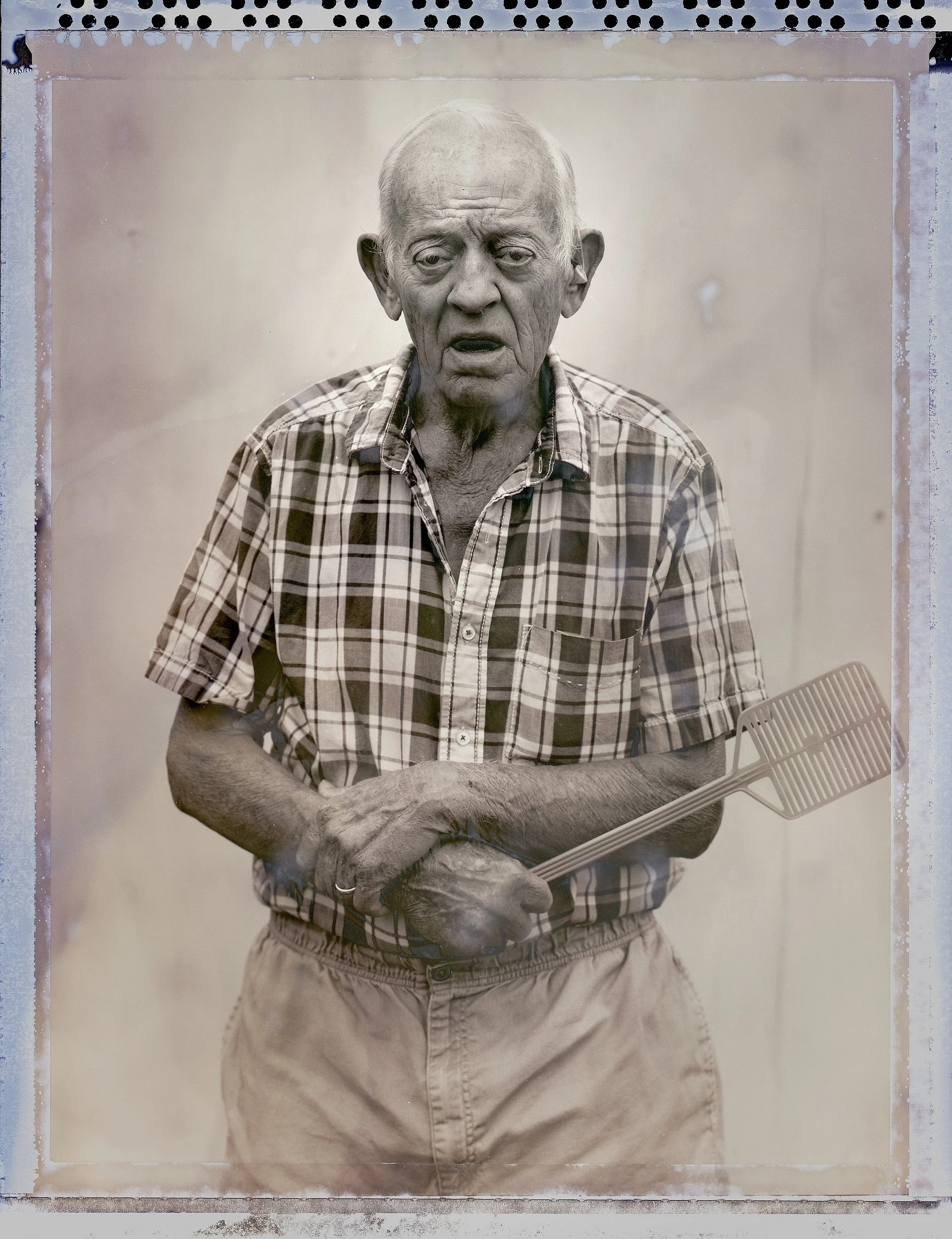 a polaroid film portrait of an older man in a plaid shirt holding a plastic fly swatter, showing the distinct type 55 polaroid borders and textures of film, from One Way Out, Lord by Parker J Pfister.