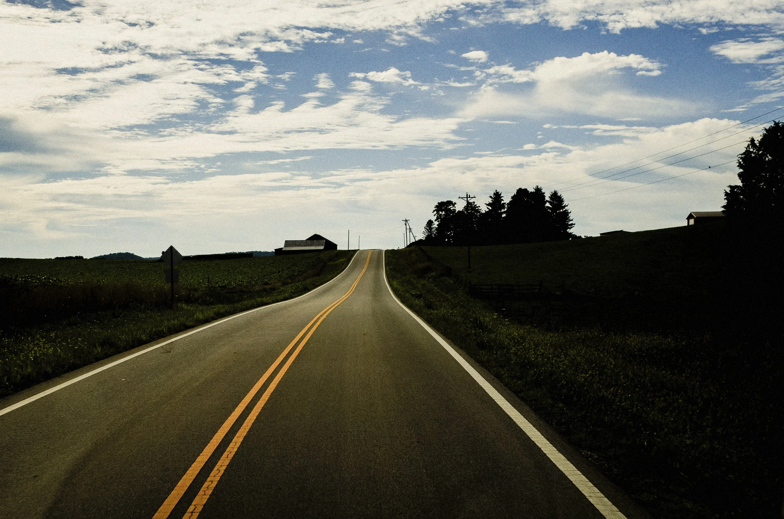 A wide color documentary photograph of an empty two-lane country highway with double yellow lines stretching into the distance through rural Ohio fields under a vast blue sky with white clouds, from One Way Out, Lord by Parker J Pfister.