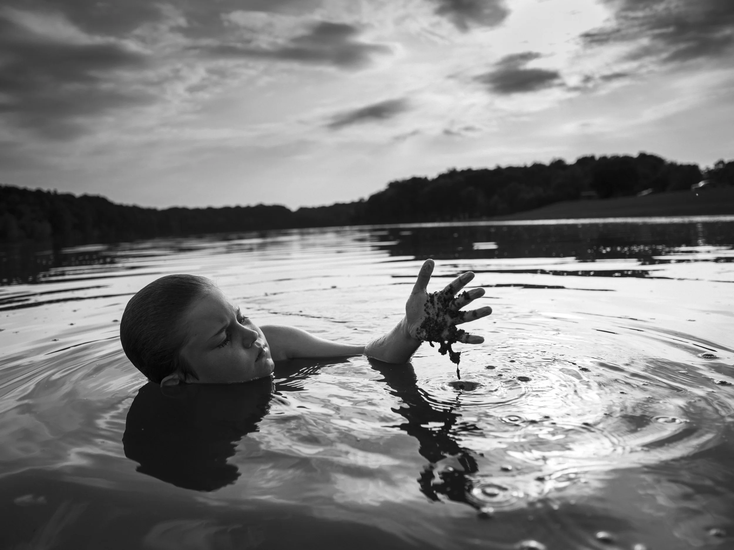 A striking, low-angle black and white photograph of a boy wading in a lake, holding a handful of lake weeds out of the water against a dramatic, cloudy sky, from One Way Out, Lord by Parker J Pfister.