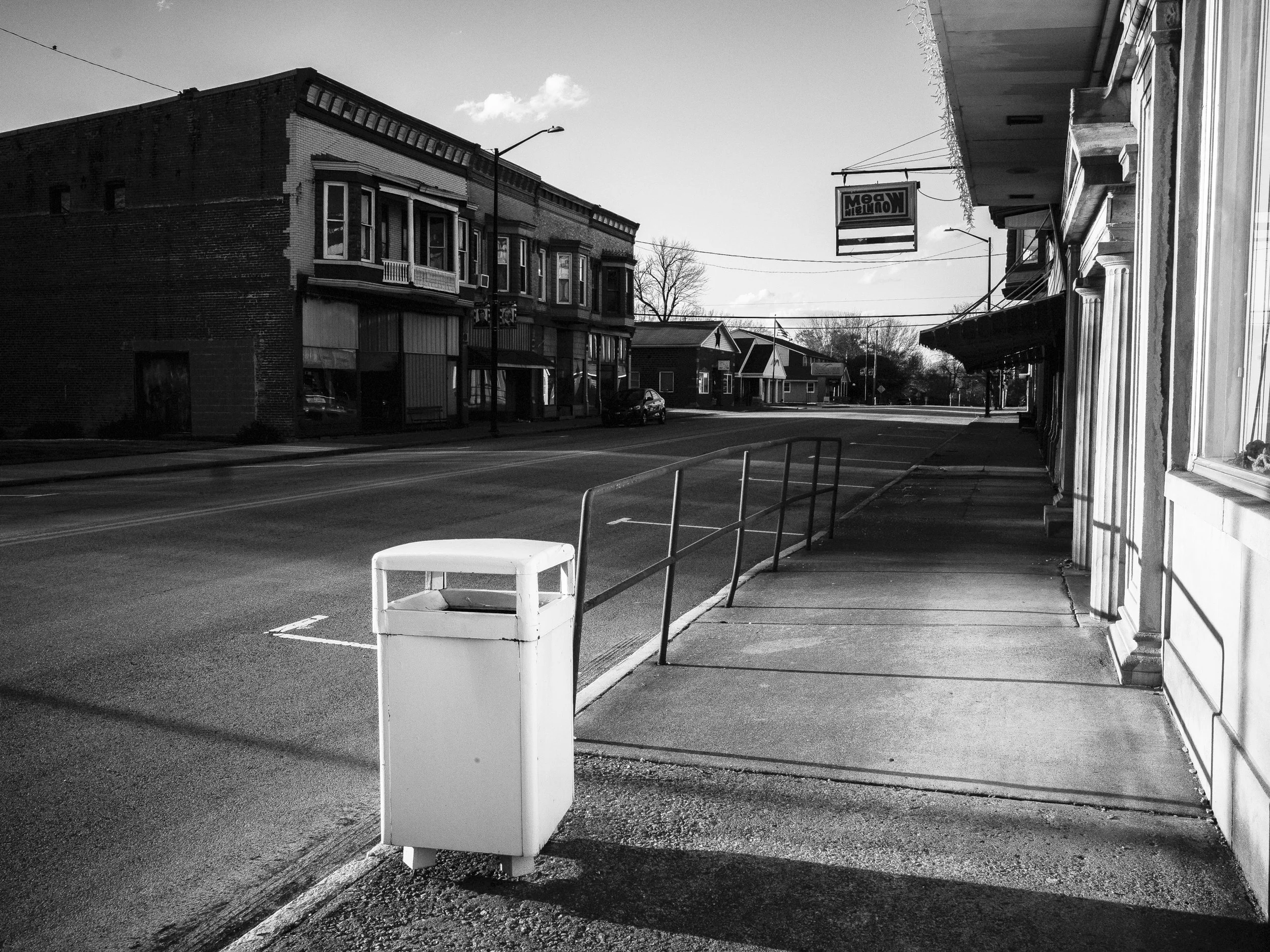 A crisp black and white environmental photograph looking down an empty commercial street and sidewalk with a vintage hanging sign in Lynchburg, Ohio, from One Way Out, Lord by Parker J Pfister.