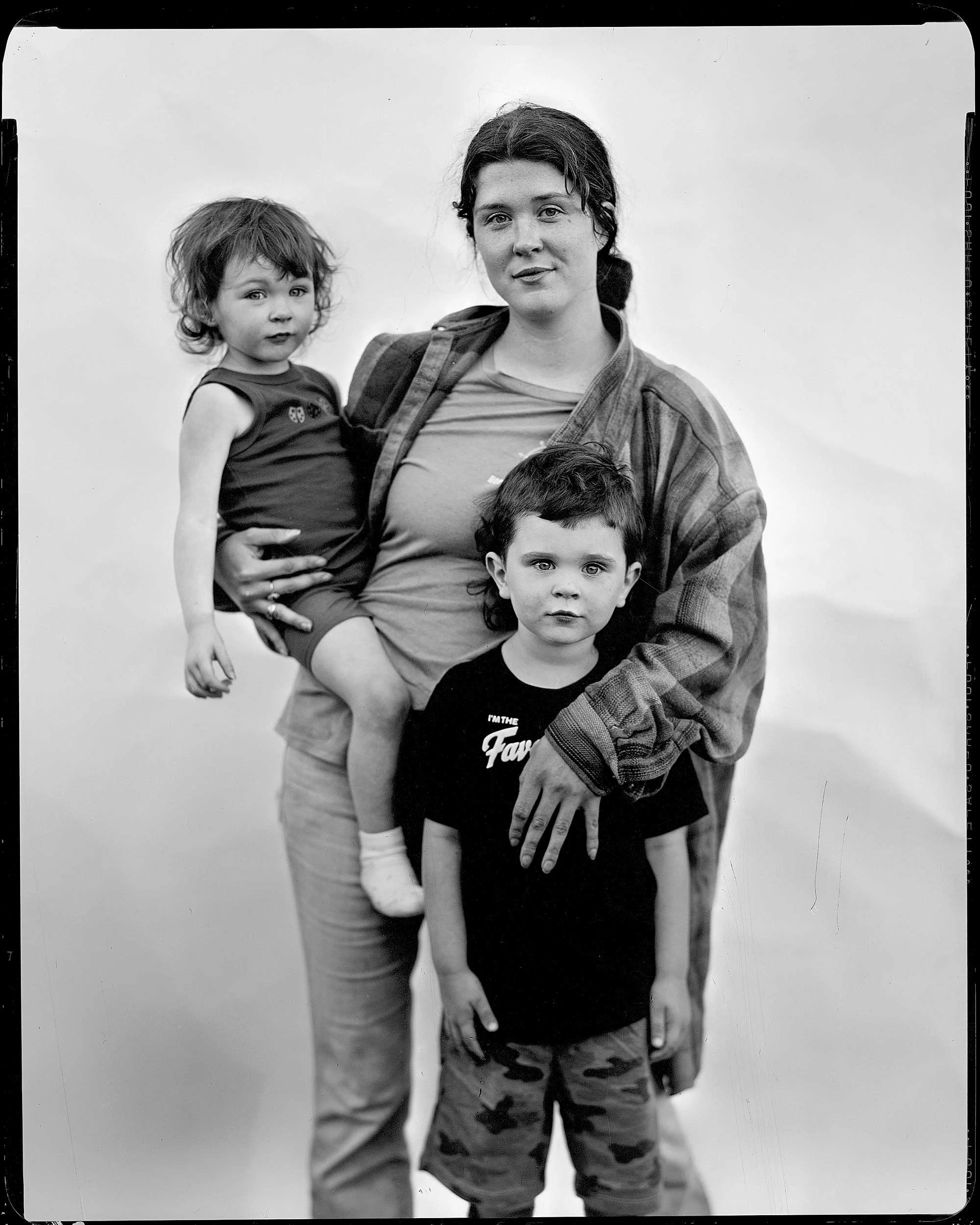 An 8x10 large format black and white paper negative portrait of a young mountain mother standing with her two young children—holding a toddler in her arms while her young son stands in front wearing a black graphic t-shirt