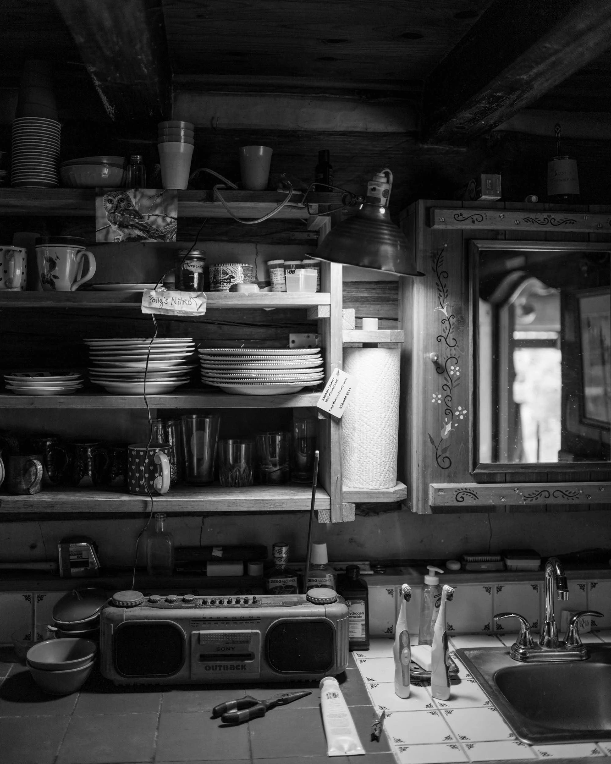 Black and white documentary photograph of a rustic kitchen counter in a Western North Carolina home, featuring an old portable radio, kitchen sink, open shelving with plates and mugs, and a small decorative mirror.