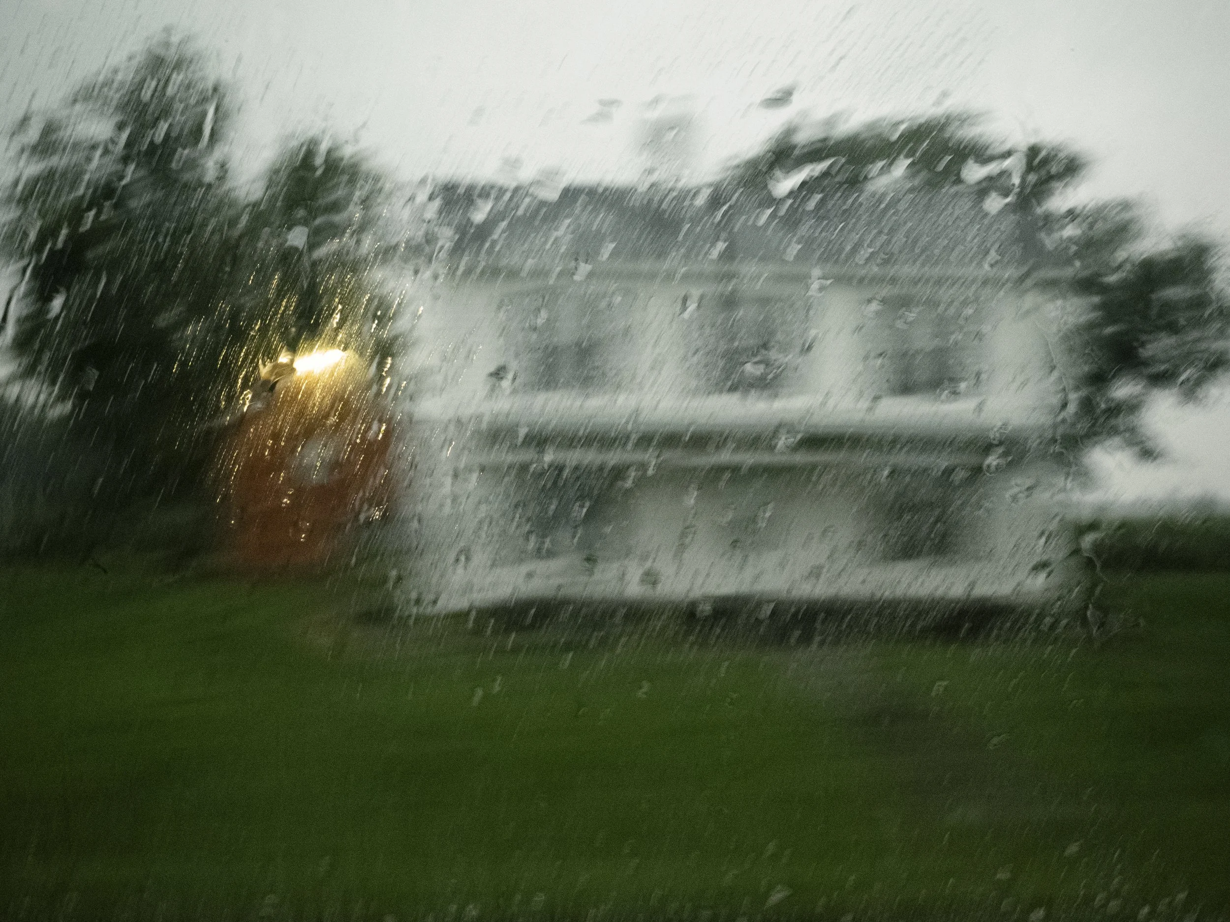 A moody, abstract color photograph capturing a two-story white house blurred through a heavy downpour and rain-streaked glass, with a warm light glowing from a window, from the One Way Out, Lord series by Parker J Pfister.