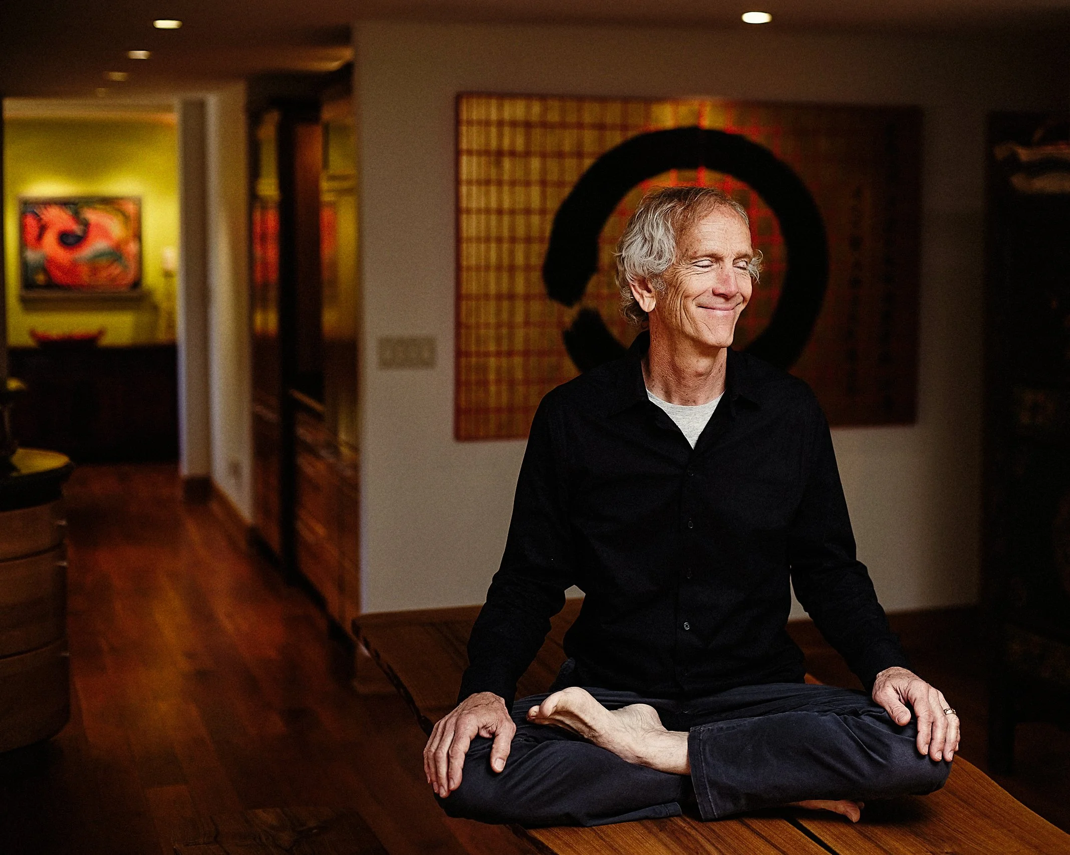 Warm, relaxed indoor color portrait of a man sitting cross-legged on a wooden surface in a meditative pose, smiling gently in a softly lit interior space with art pieces in the background.