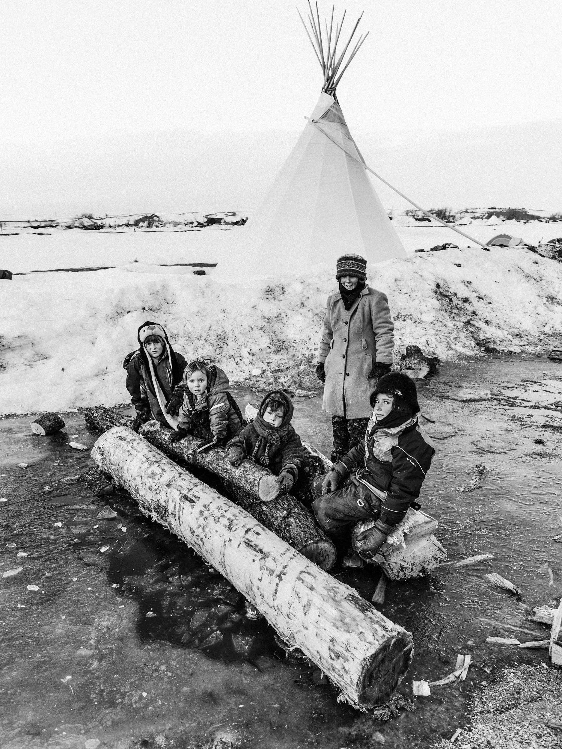 Black and white documentary photograph of four young children bundled in winter coats sitting together on large fallen logs in the snow. A woman stands just behind them, and a traditional white tipi rises into the pale sky in the background.