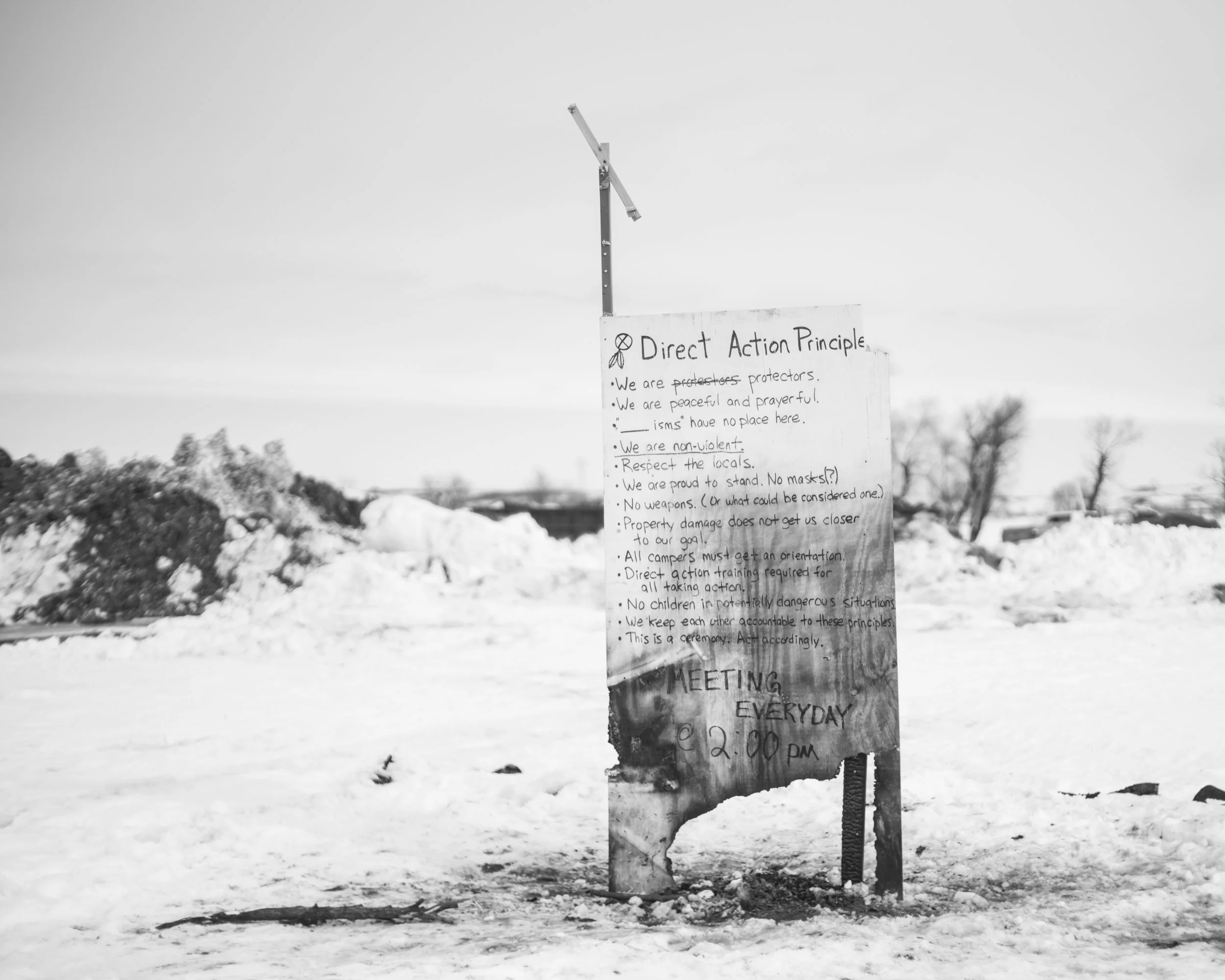 Black and white photograph of a tall wooden sign planted in a snow-covered field at the Standing Rock camp. The sign lists handwritten guidelines titled "Direct Action Principle" and notes "Meeting Everyday 2:00 PM" at the bottom.