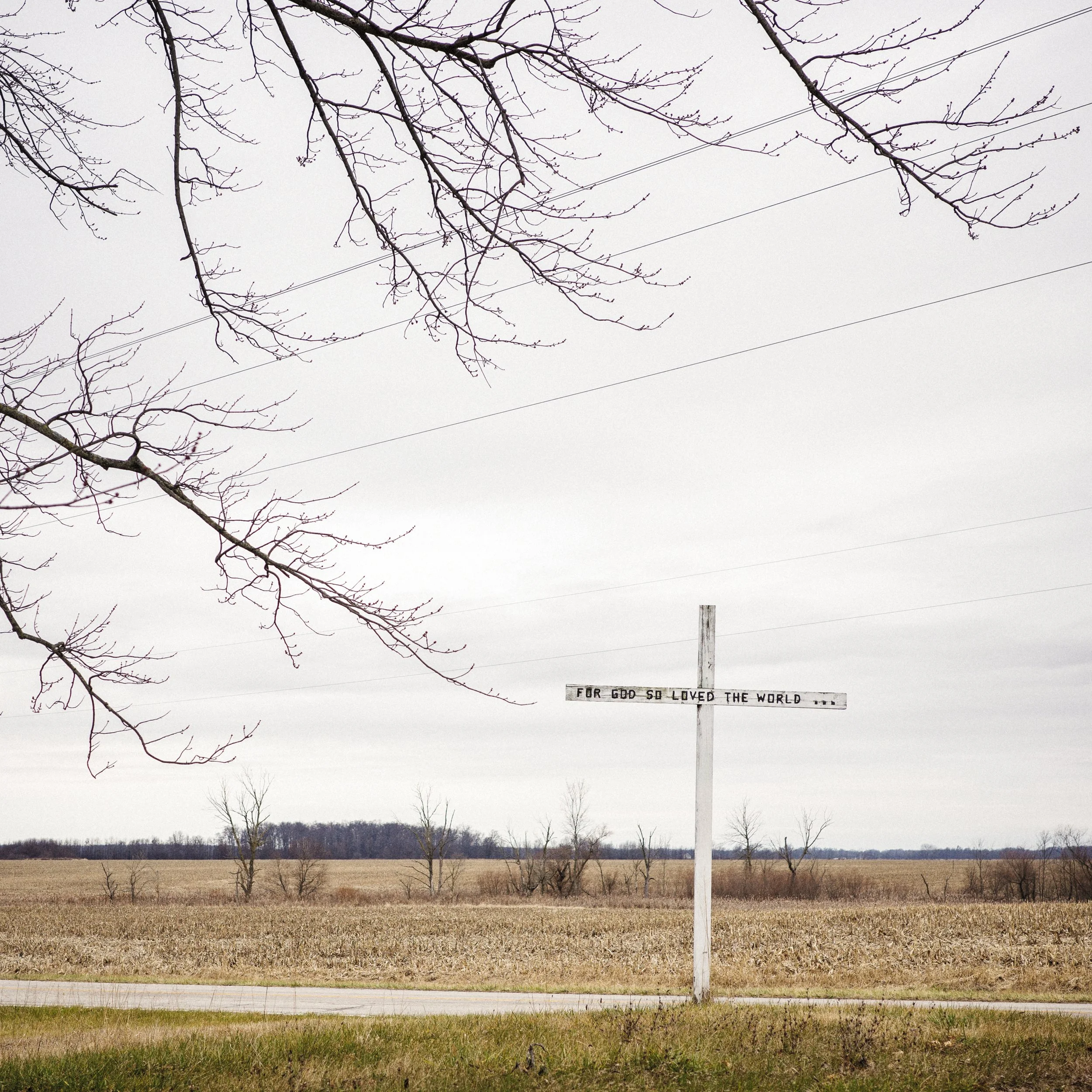 A color documentary photograph of a tall white Christian cross bearing the inscription "FOR GOD SO LOVED THE WORLD" standing in a vast, flat Ohio field beneath bare tree branches, from One Way Out, Lord by Parker J Pfister.