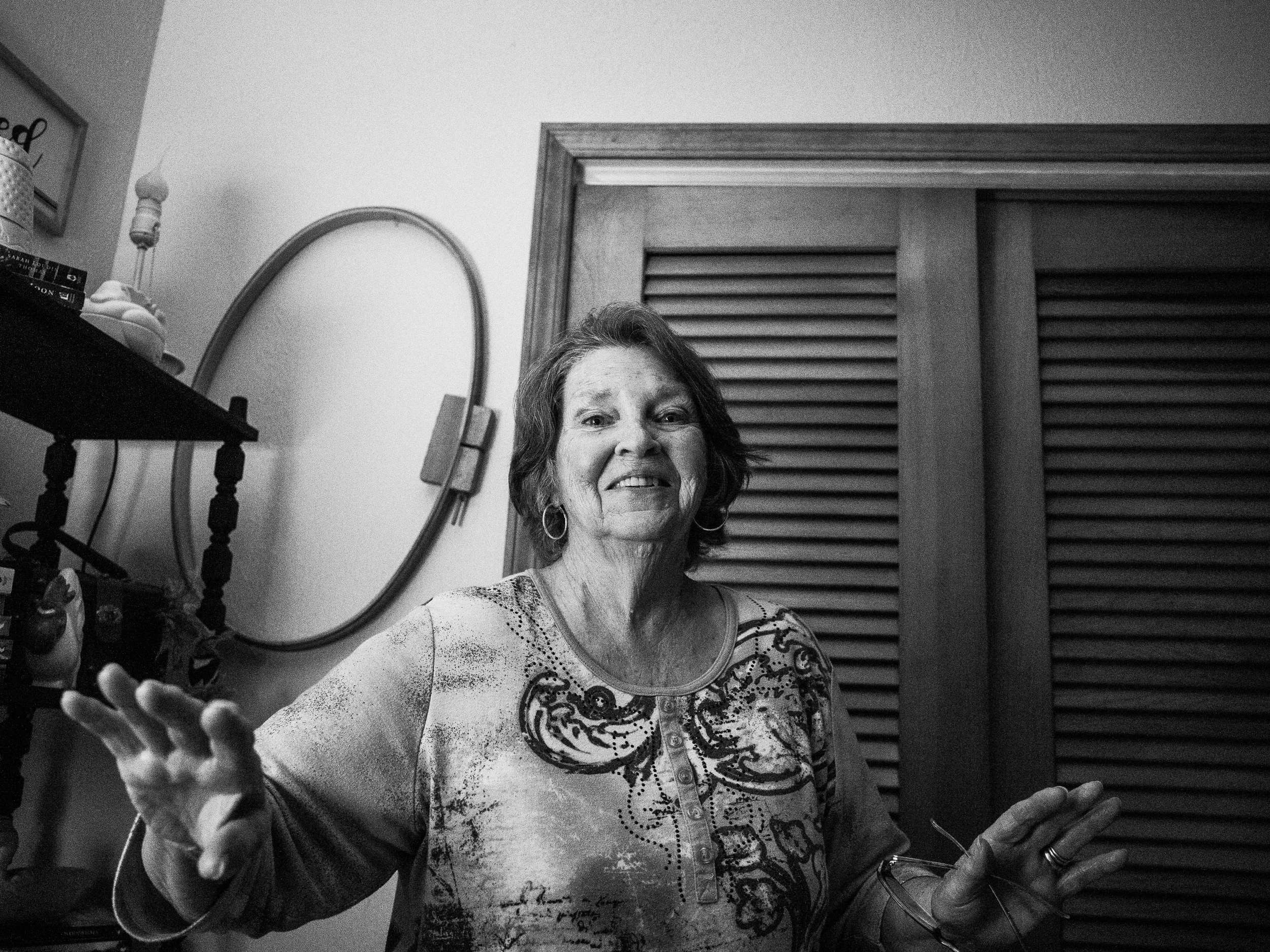 An intimate black and white indoor portrait of an older woman smiling and gesturing with her hands, with a wooden slatted door in the background, from the One Way Out, Lord series by Parker J Pfister.