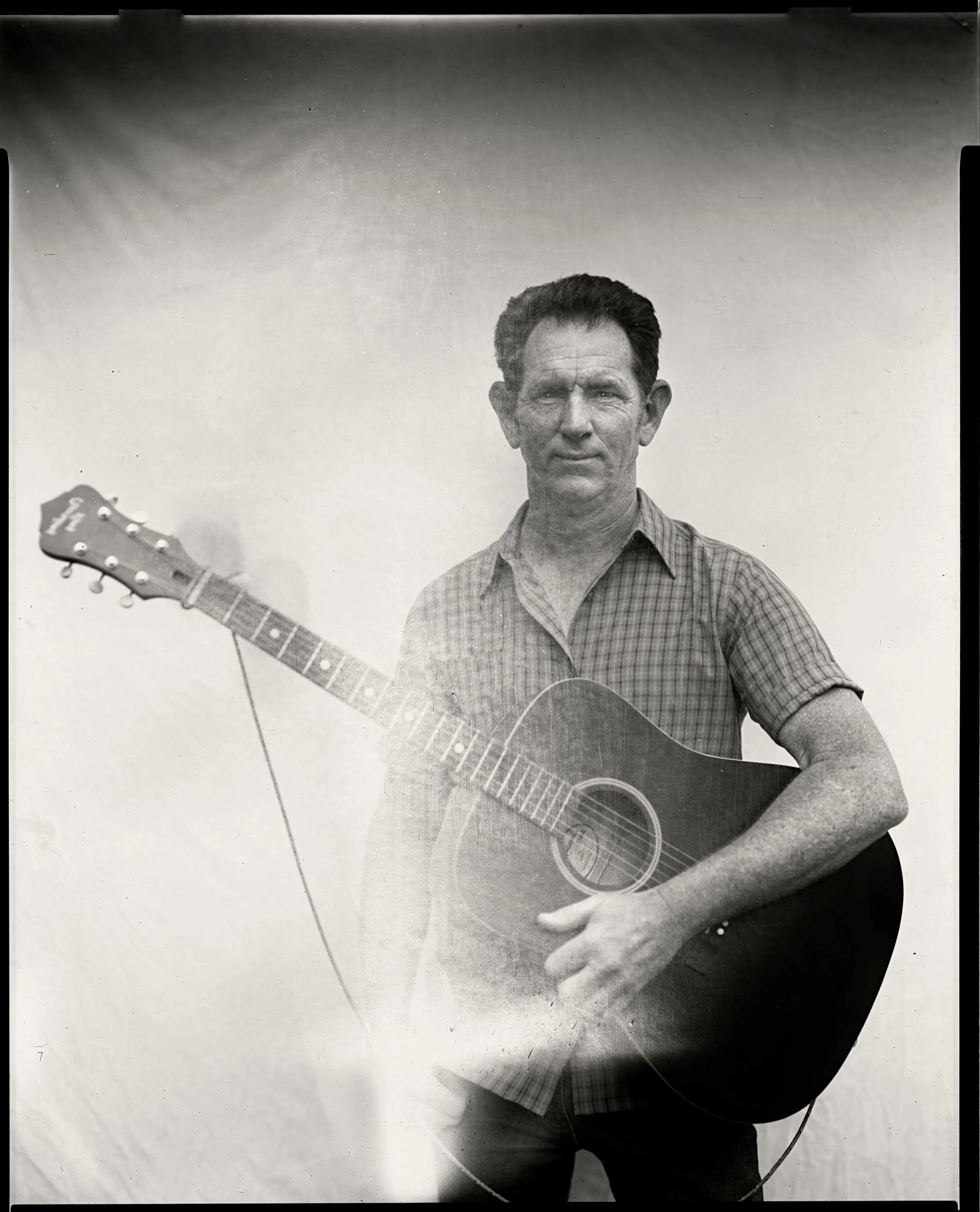 An 8x10 large format black and white paper negative portrait of a man in a plaid shirt holding an acoustic guitar, captured at the Bluff Mountain Music Festival in Hot Springs, North Carolina.