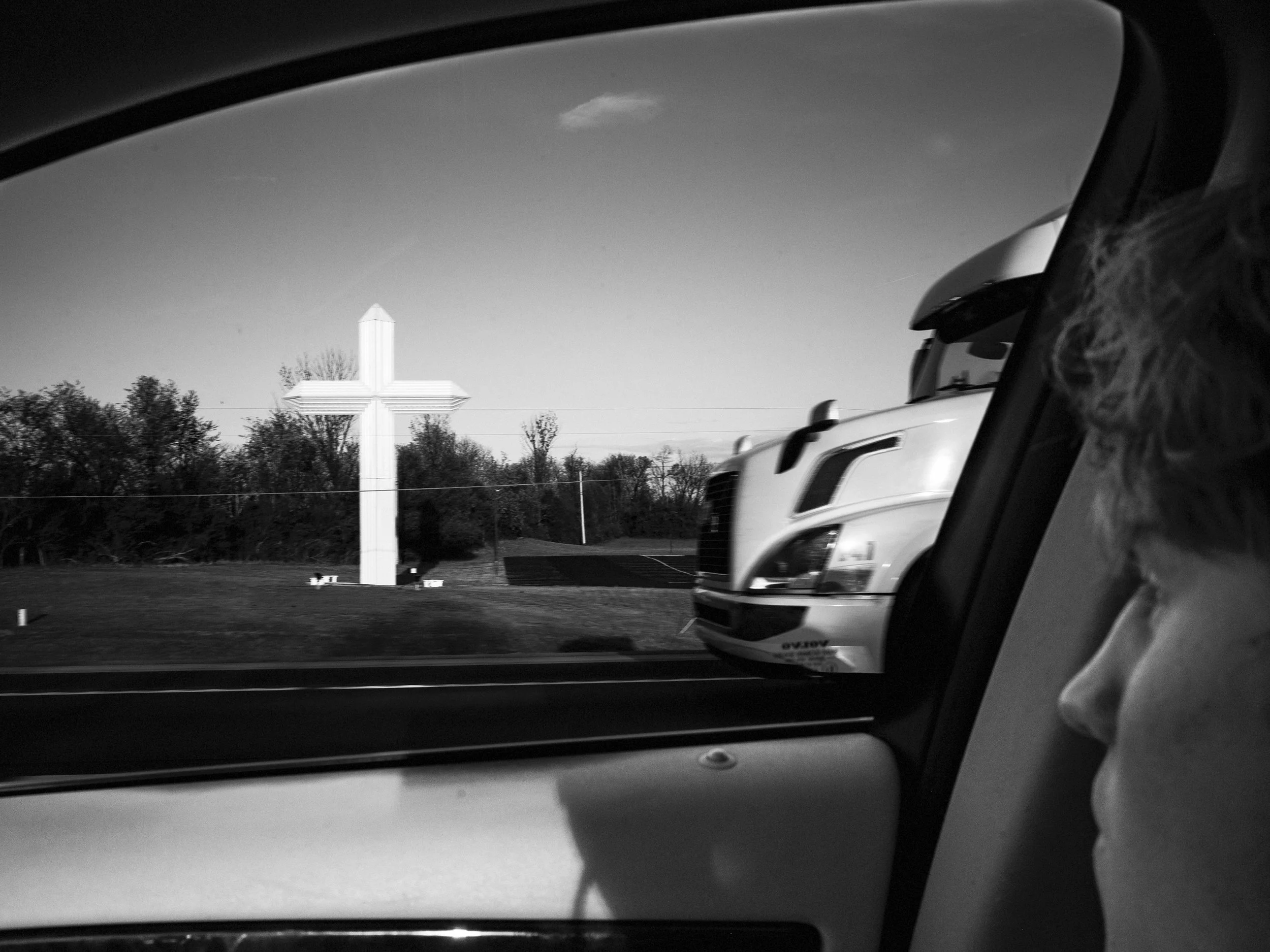 A black and white documentary photograph taken from inside a car window, framing a giant white roadside Christian cross and a passing semi-truck, with a passenger's profile in the foreground, from One Way Out, Lord by Parker J Pfister.