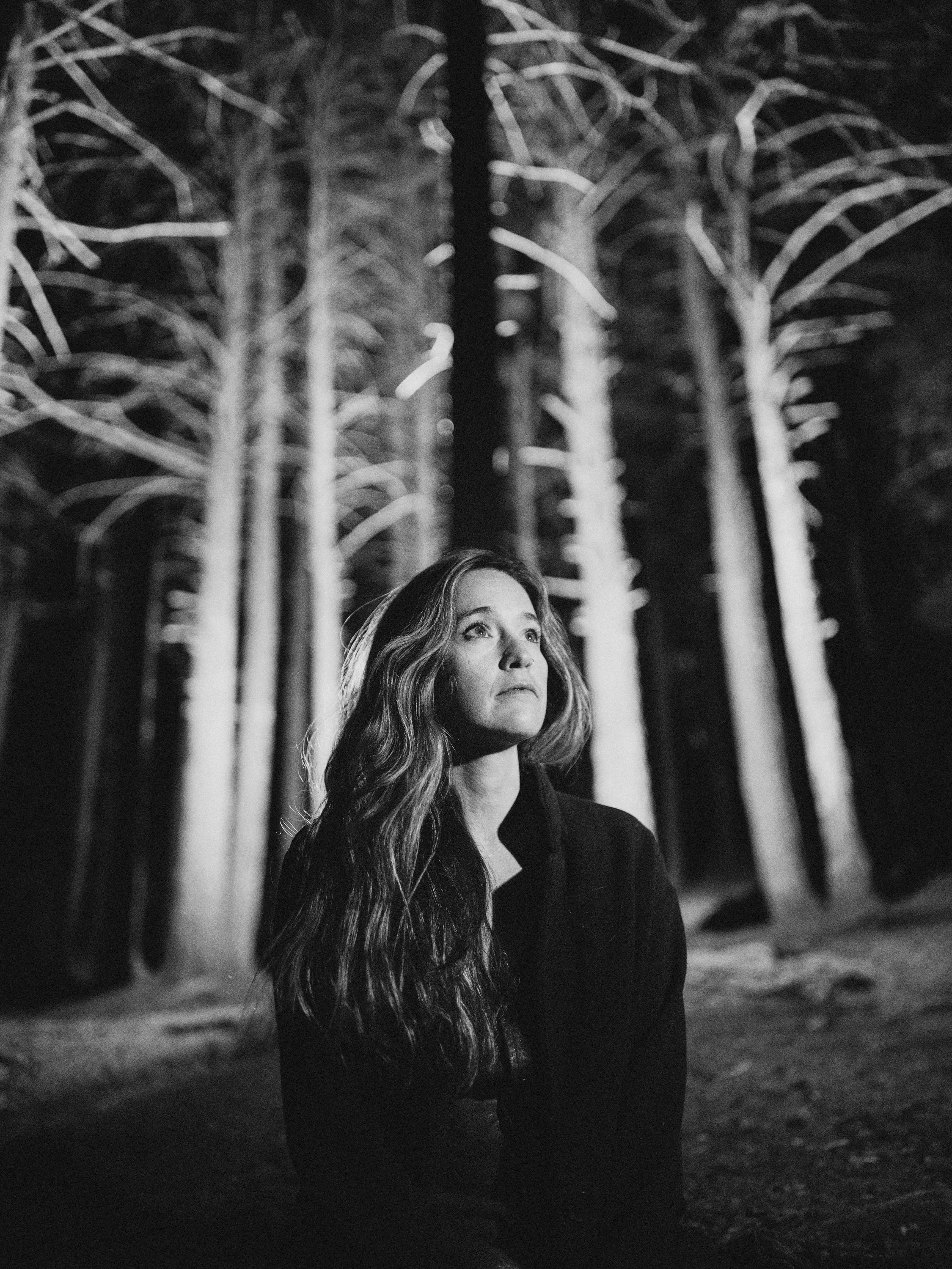 Black and white portrait of singer-songwriter Lindsey Wall, frontwoman of Western North Carolina band Blue Ridge Betty, standing in a pine forest for the album Demons, photographed by Parker J. Pfister.