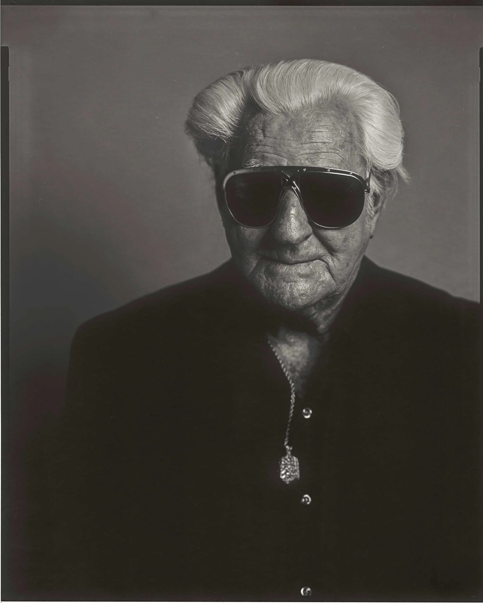 An 8x10 large format black and white paper negative portrait of an older man with swept-back white hair wearing dark aviator sunglasses and a button-down shirt with a necklace, captured at the old Depot in Marshall, North Carolina.