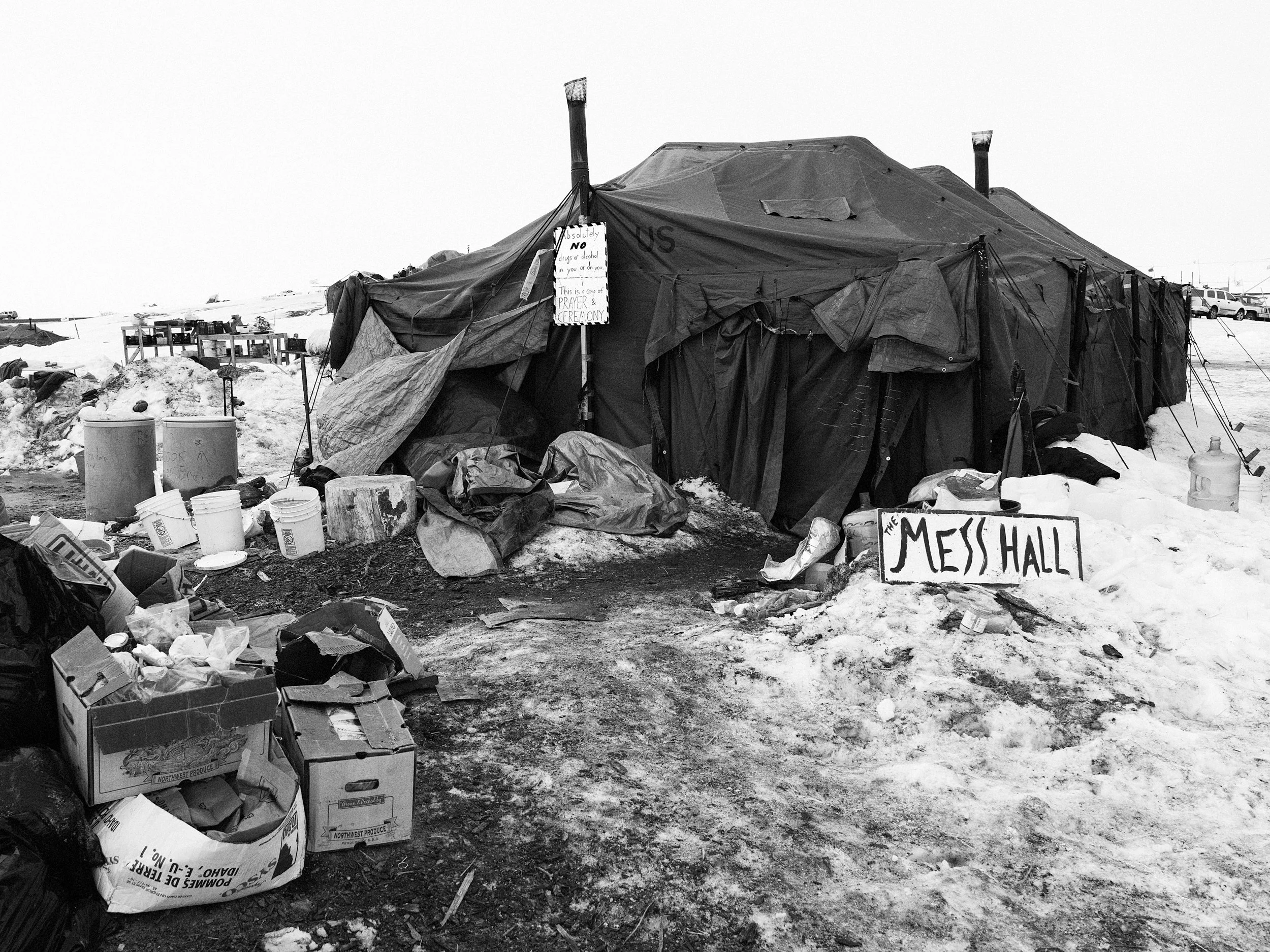 Black and white documentary photograph of a snow-covered section of the Standing Rock camp, featuring cardboard supply boxes stacked on the left and a handmade wooden sign on the right that reads "MESS HALL."