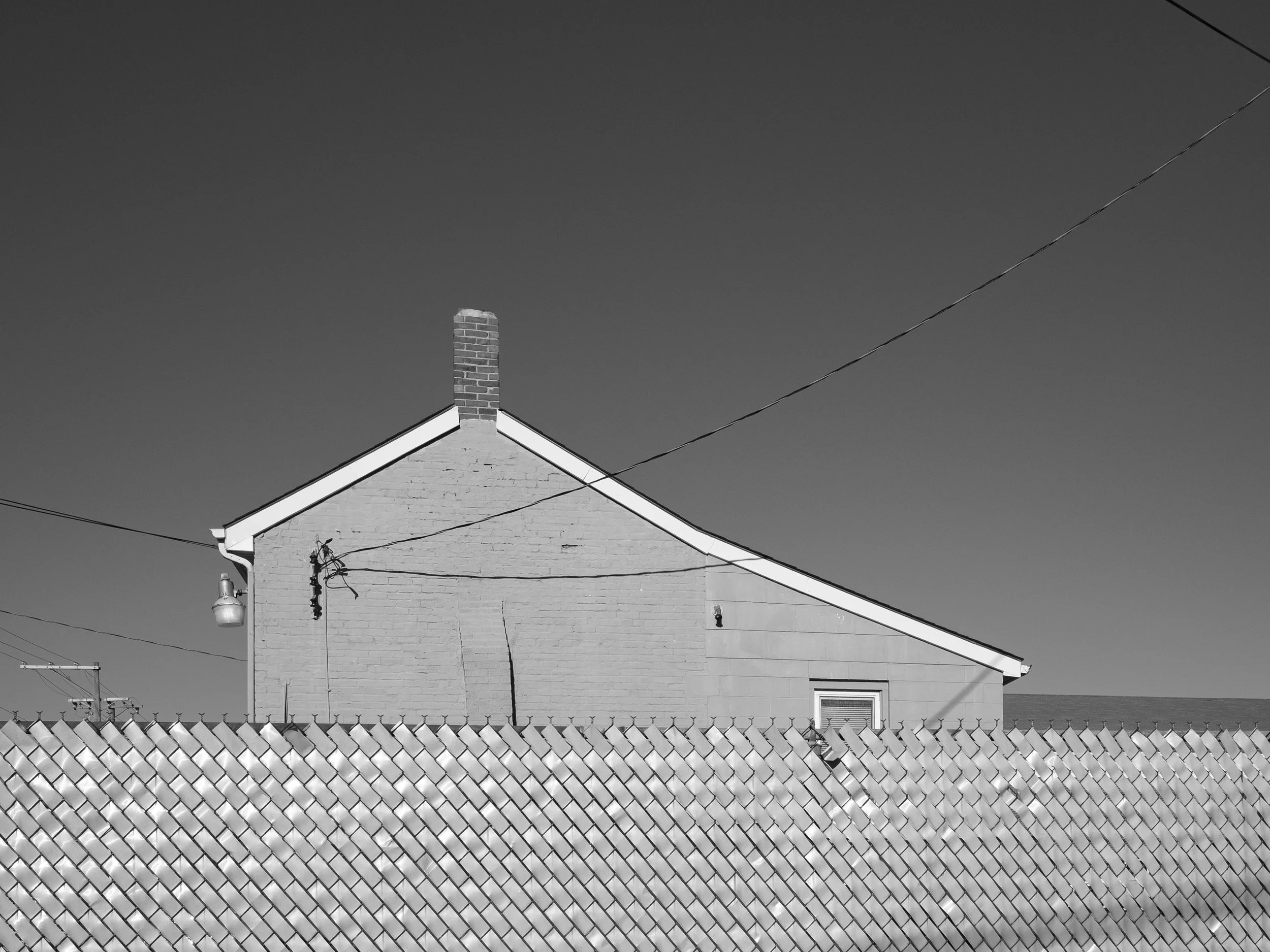 A stark black and white documentary photograph of a house roofline and chimney framed above a long chainlink fence with privacy slats, from One Way Out, Lord by Parker J Pfister.