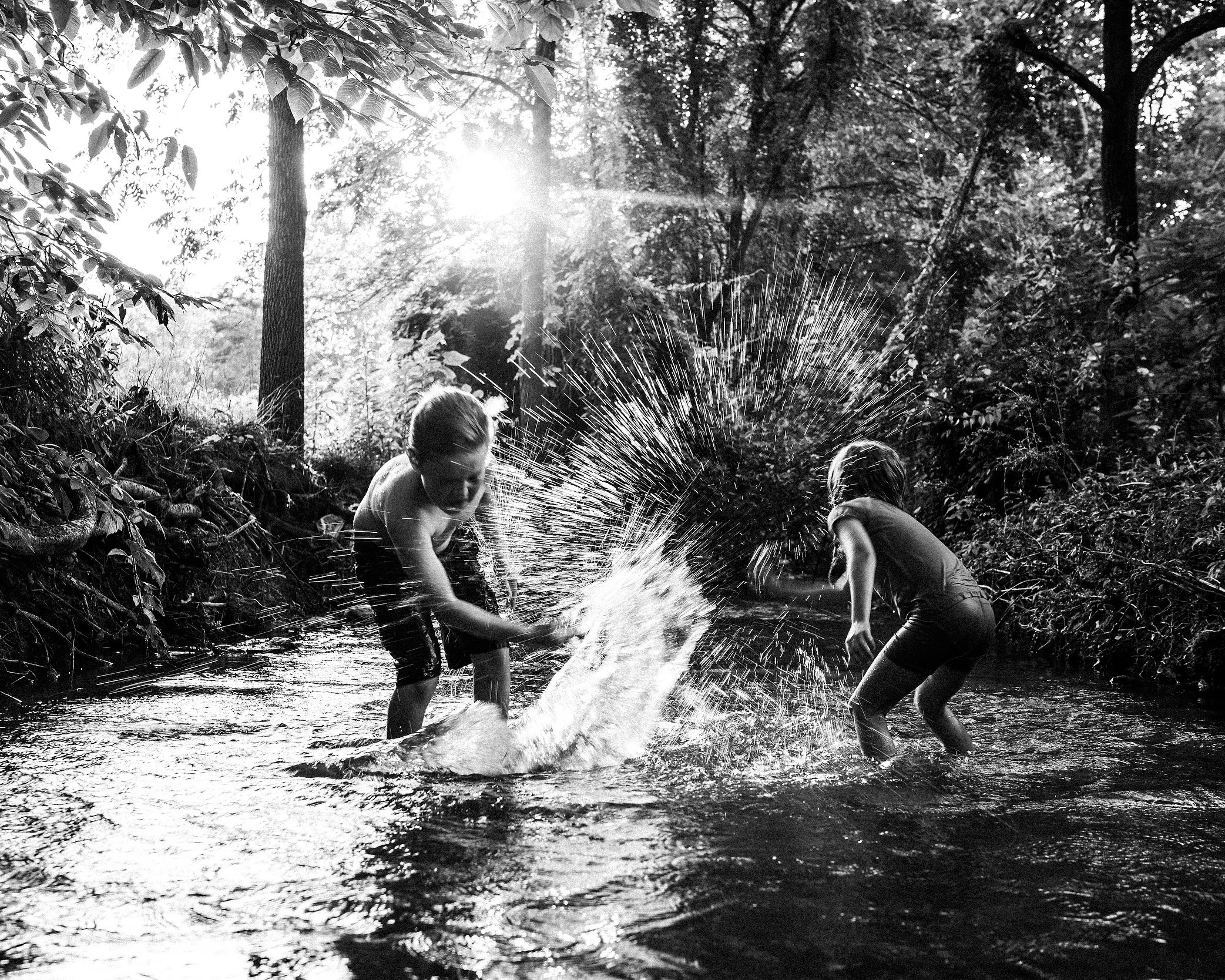Black-and-white action photograph of two children splashing playfully in a sun-dappled mountain stream, surrounded by lush forest trees, with bright water droplets frozen mid-air in the sunlight.