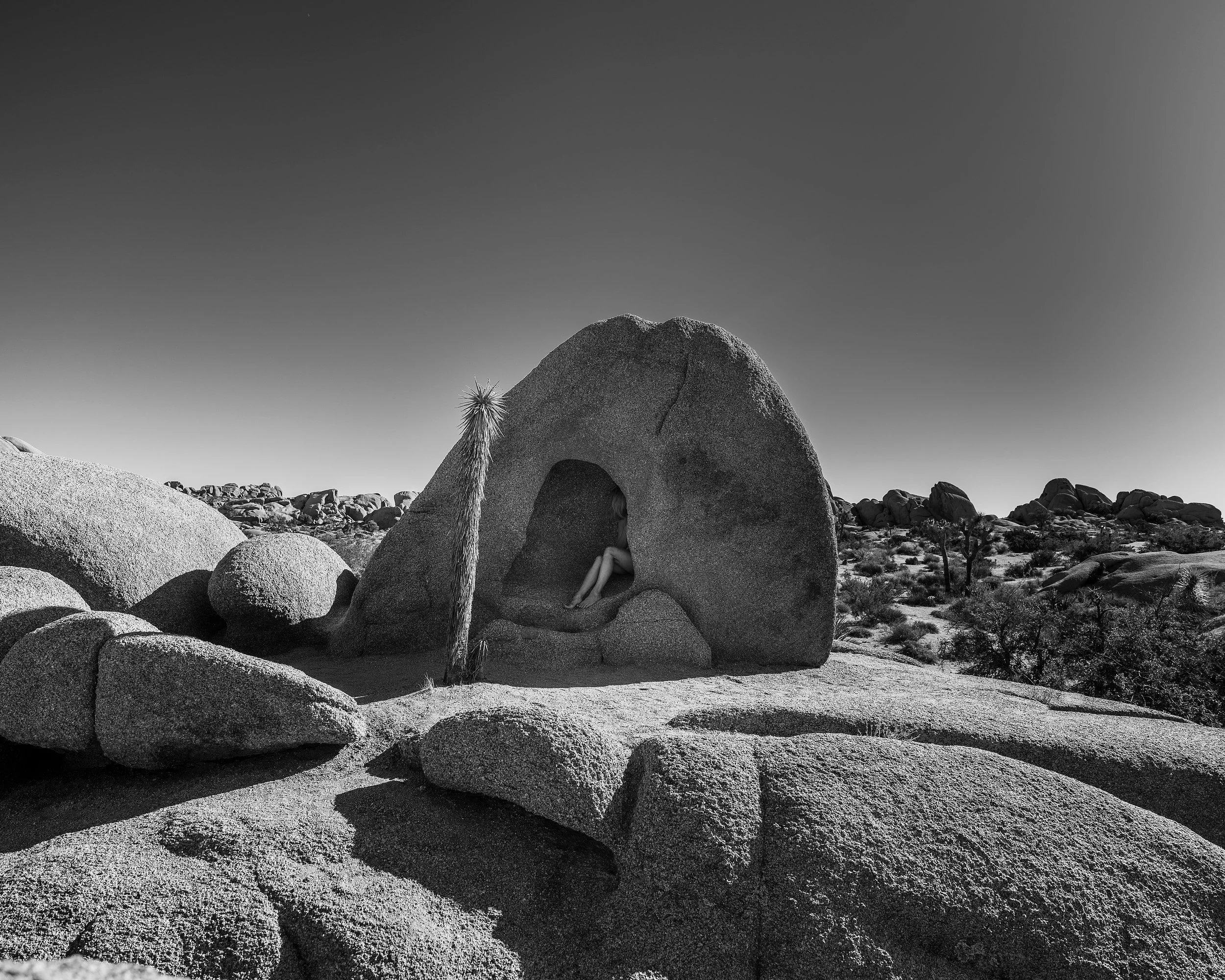 Black and white fine art photograph of a nude figure nestled inside a small, hollowed-out opening of a massive boulder in a desert landscape, with a lone joshua tree standing nearby against a clear sky.
