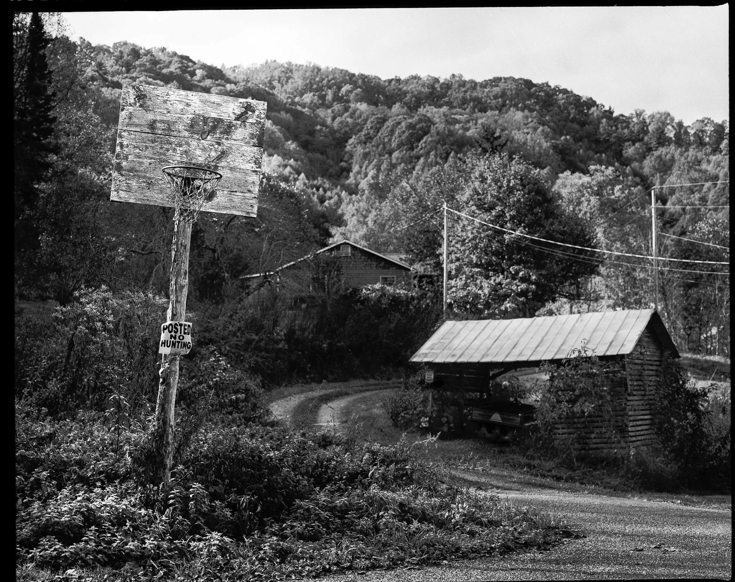 A high-contrast black and white documentary photograph of a weathered, homemade wooden basketball hoop with a "POSTED NO HUNTING" sign nailed to the post, standing at the edge of a gravel road leading into the wooded hills of Appalachia.