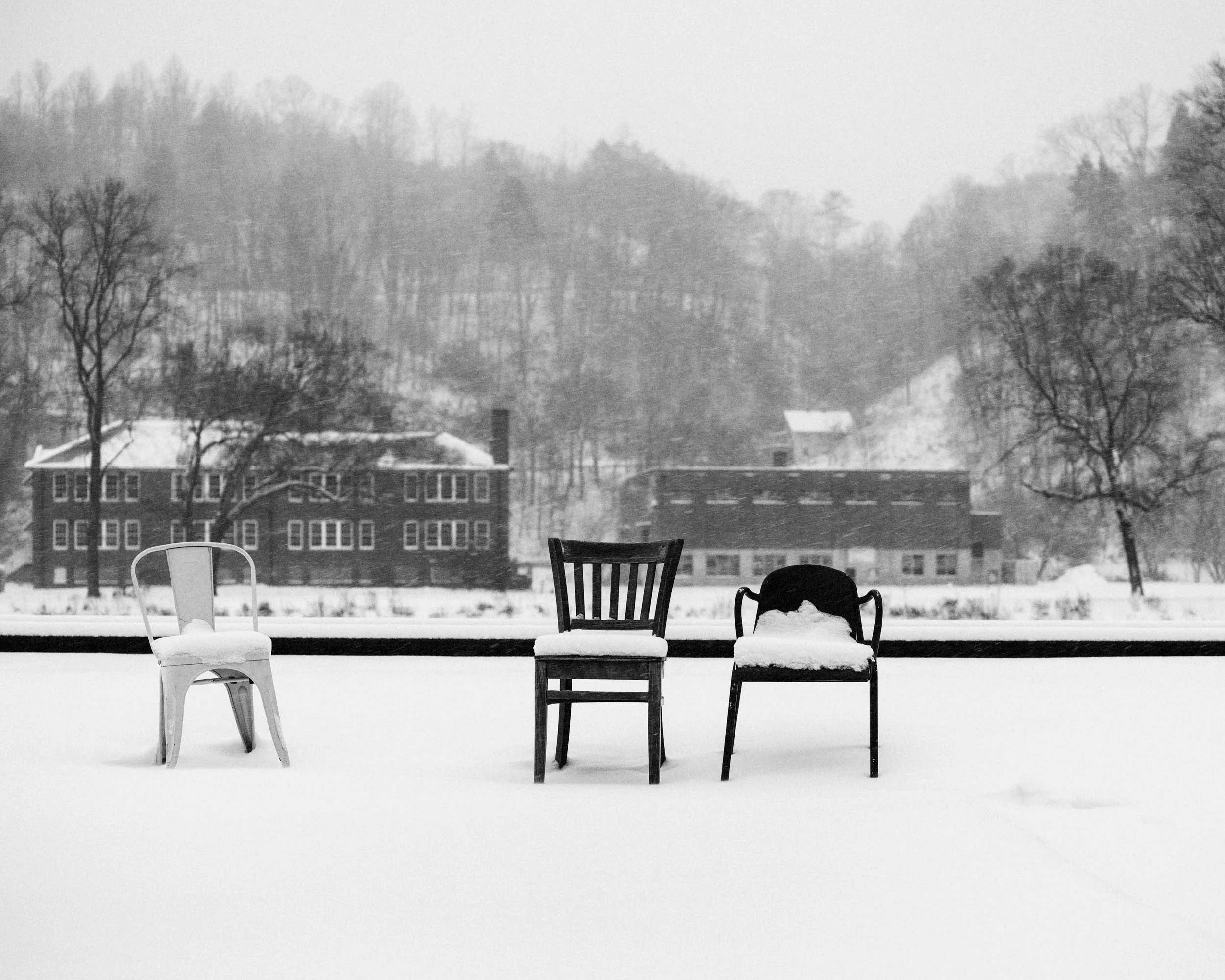 A quiet and poetic black and white documentary photograph showing three empty, mismatched chairs lined up in deep snow, looking out toward a historic brick school building and mist-covered winter mountains in Madison County, North Carolina.