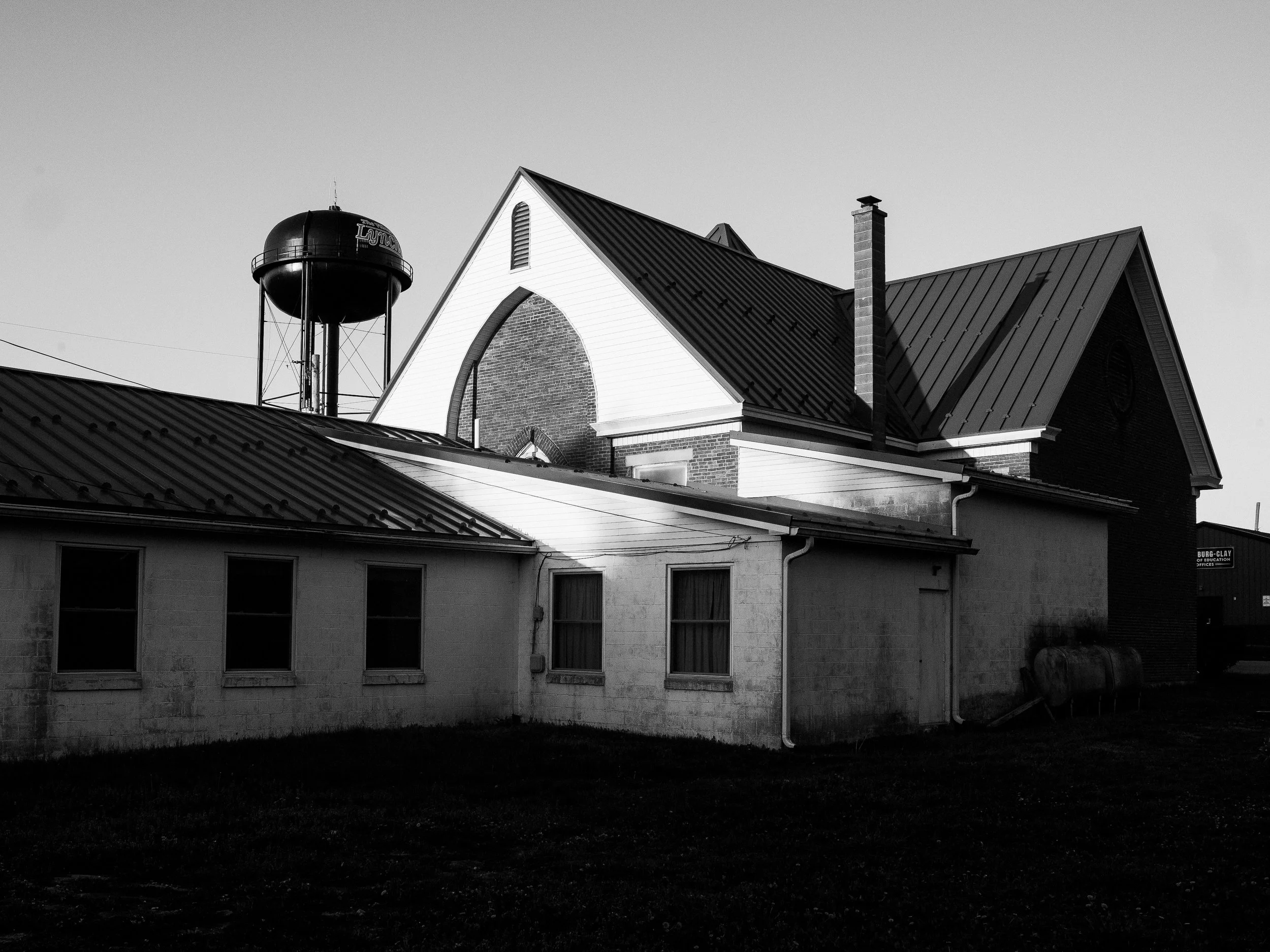 A high-contrast black and white documentary photograph of an old church building with a large water tower looming behind the roofline in Lynchburg, Ohio, from One Way Out, Lord by Parker J Pfister.