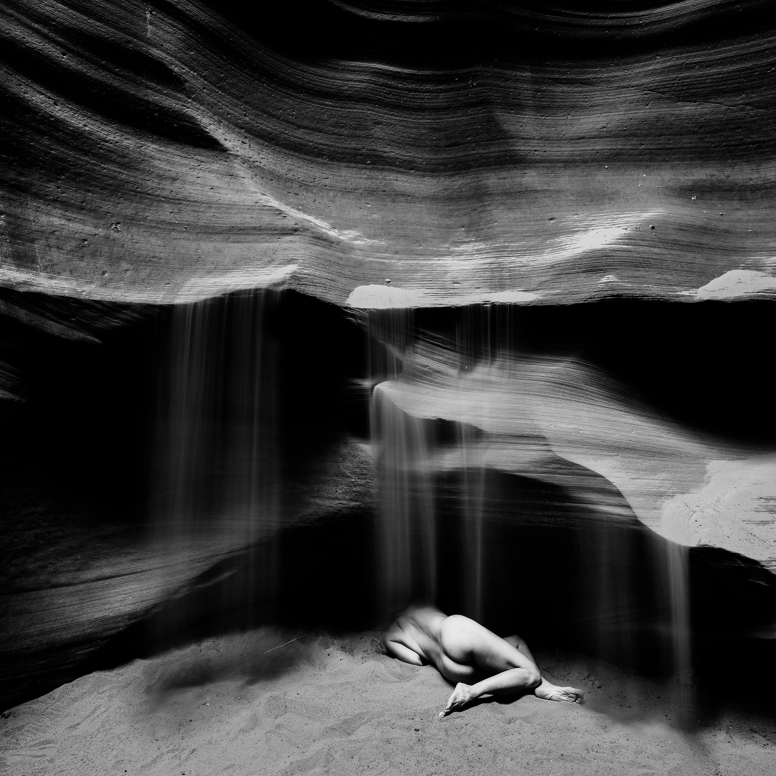 Long exposure black and white fine art photograph capturing sand cascading down smooth, sweeping striated rock layers inside a dark Antelope canyon, creating a fluid, abstract motion effect.