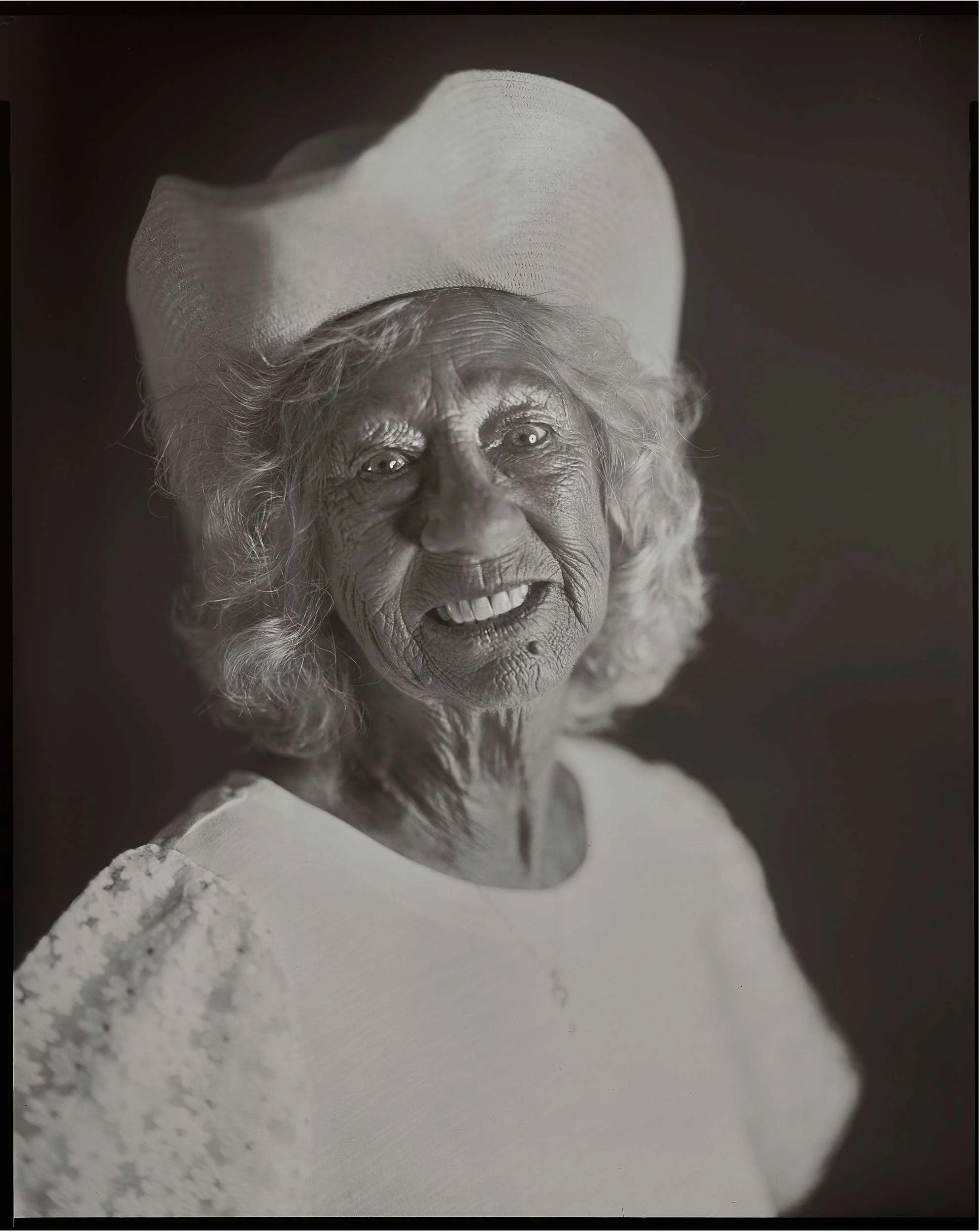 An 8x10 large format black and white paper negative portrait of a smiling elderly Appalachian woman wearing a light-colored hat and a lace-accented white dress, photographed at the old Depot in Marshall, North Carolina.