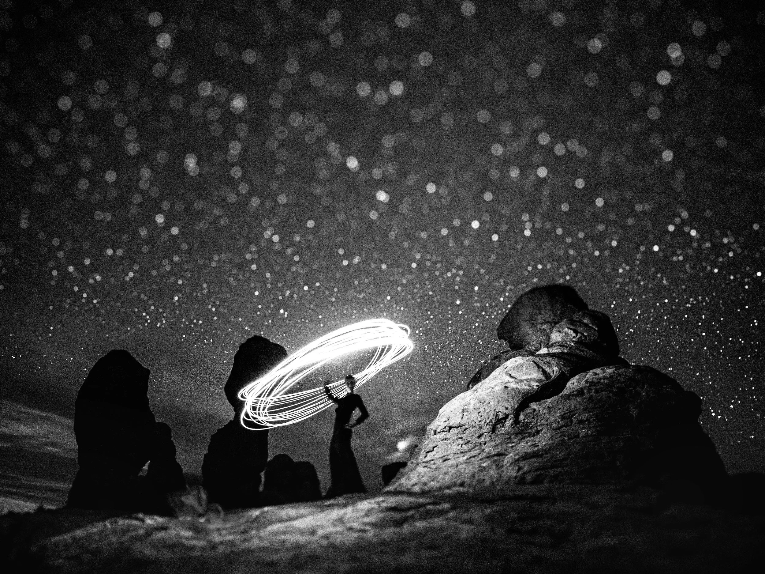 Long exposure black and white fine art photograph capturing a silhouette of a person spinning a glowing lasso of light above their head amidst dark desert rock formations in Arches National Park, underneath a vast, dramatic starry night sky.