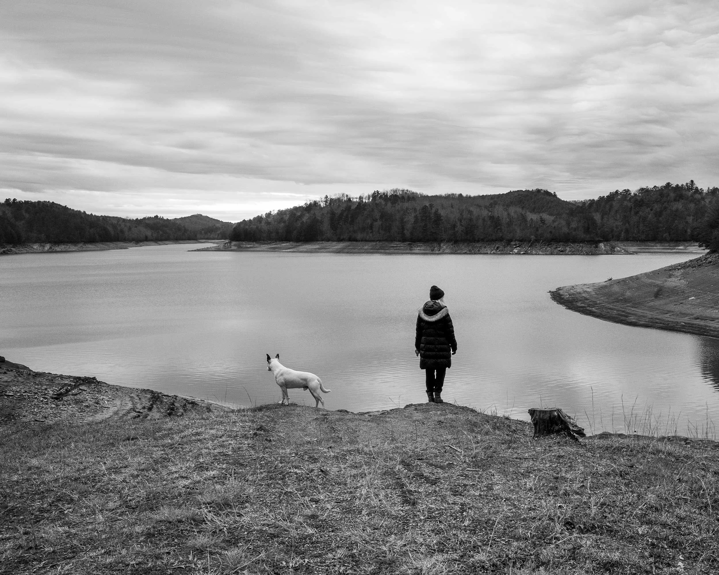 A high-contrast black and white documentary photograph of a person standing next to a white dog on a grassy bank, looking out over the quiet waters of Lake Hiwassee near Murphy, North Carolina under a dramatic cloudy sky.