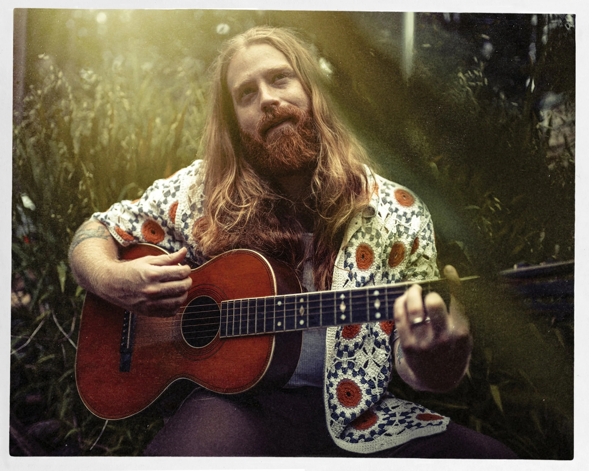 Color portrait of long-haired musician Michael Ferguson playing an acoustic guitar outdoors while wearing a patterned cardigan, photographed by Parker J. Pfister.