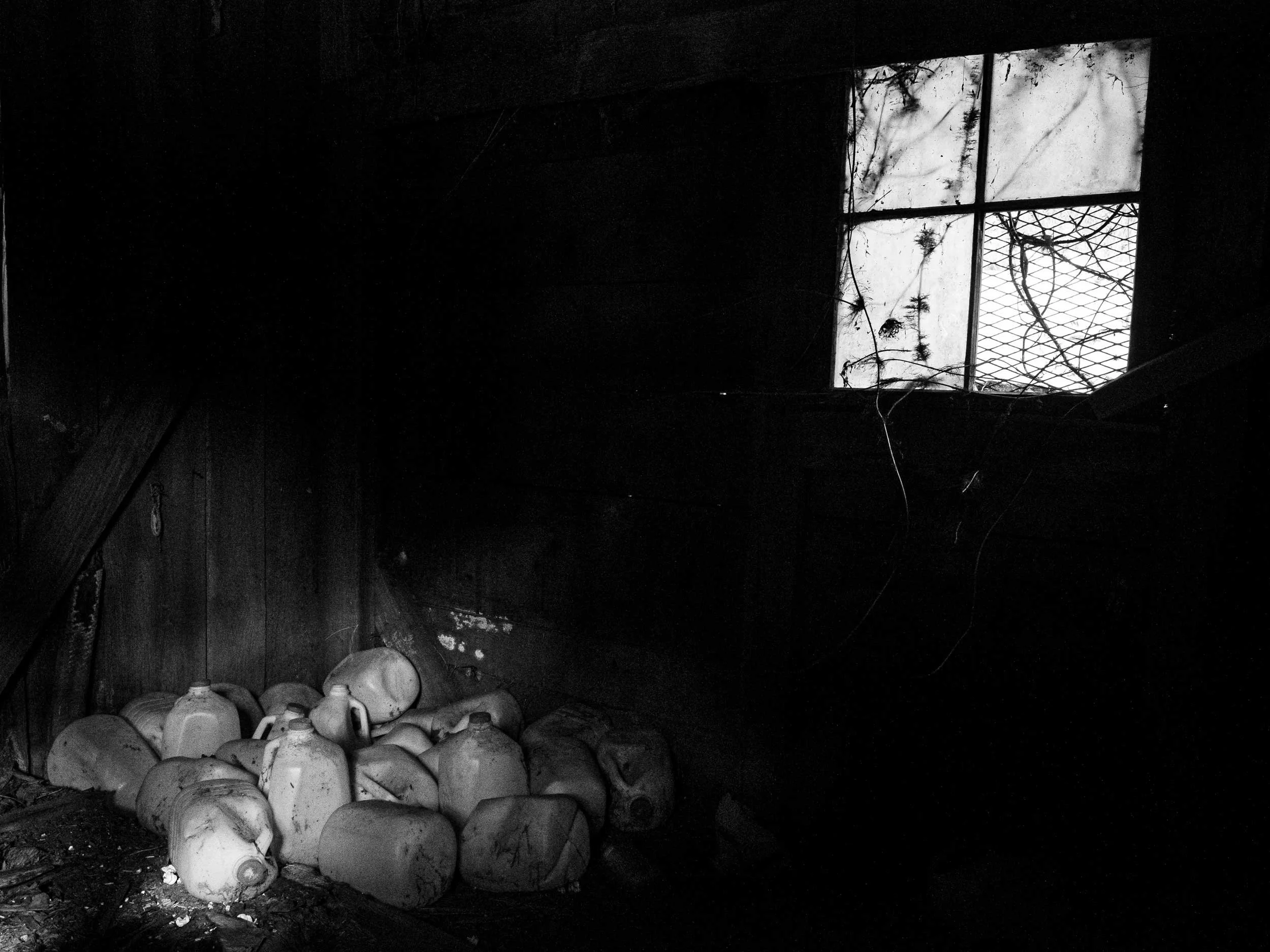 A moody, high-contrast black and white documentary photograph of a pile of old plastic milk jugs cast in shadow under a single small window with wire mesh inside a dark Appalachian barn or shed.