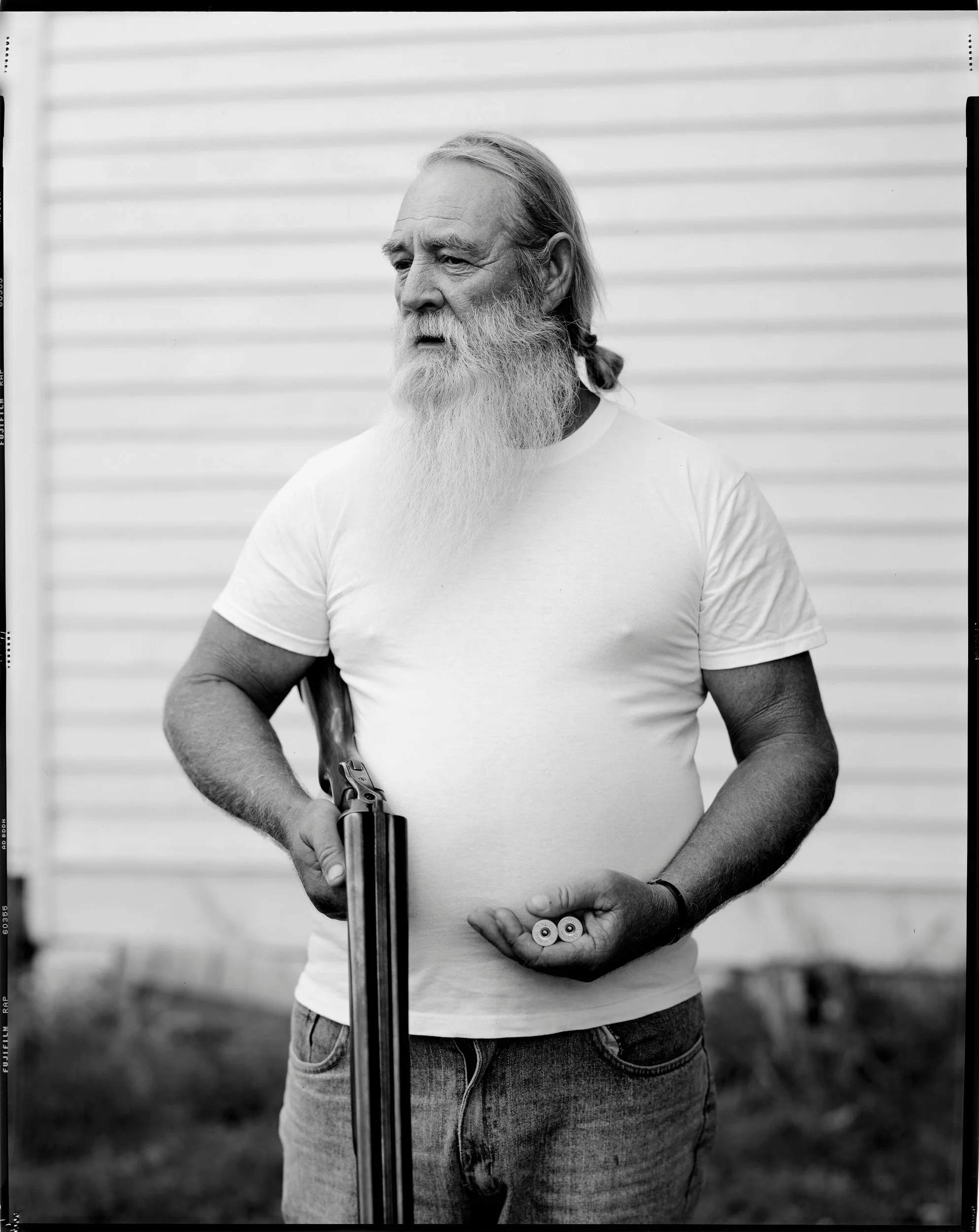 A powerful black and white environmental portrait of an older man with a long white beard holding a shotgun and two shells in his hand, from the One Way Out, Lord documentary series by Parker J Pfister.