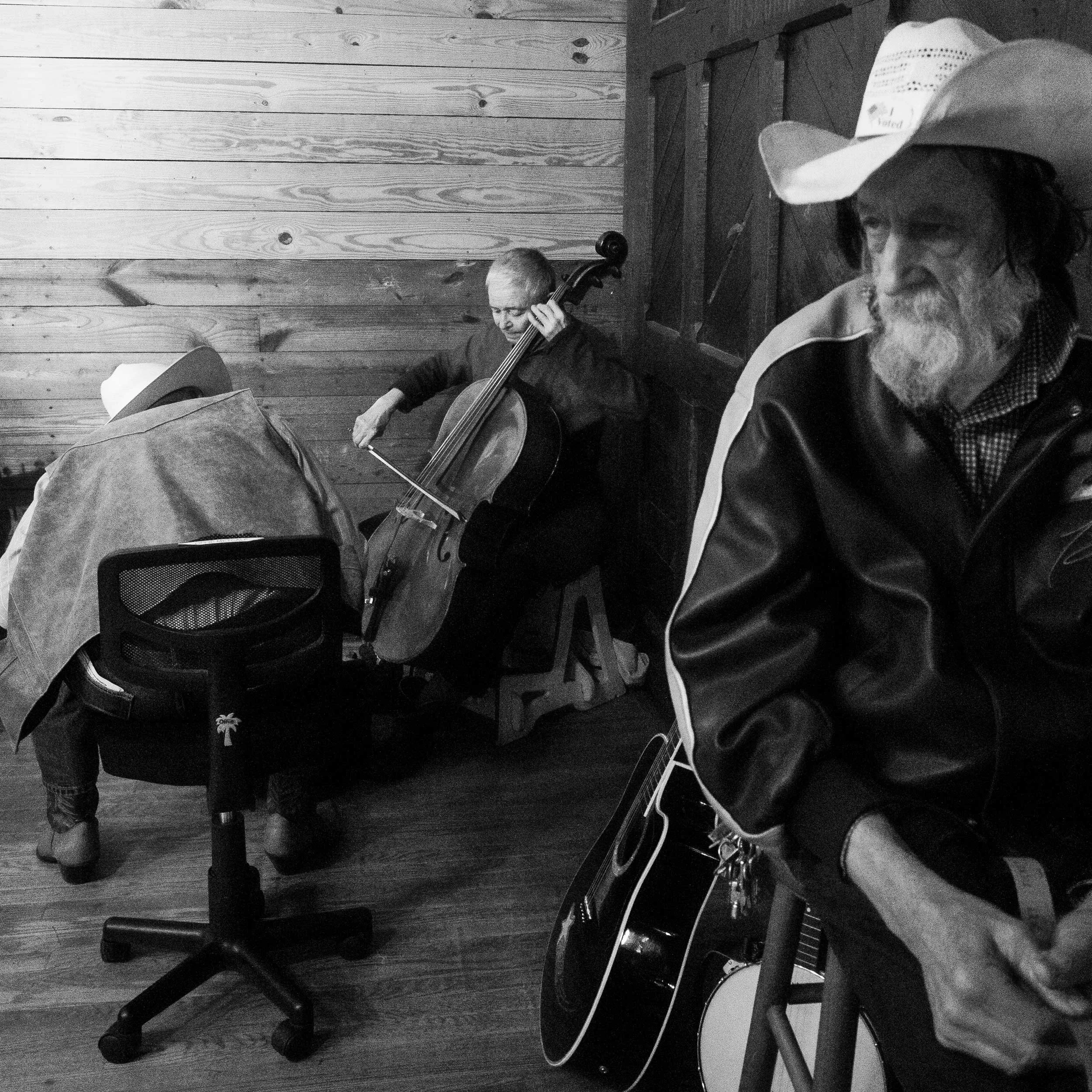 A black and white portrait capturing a group of old-time musicians inside the old Depot in Marshall, North Carolina; featuring a man in a cowboy hat in the foreground and a woman playing the cello in the background.