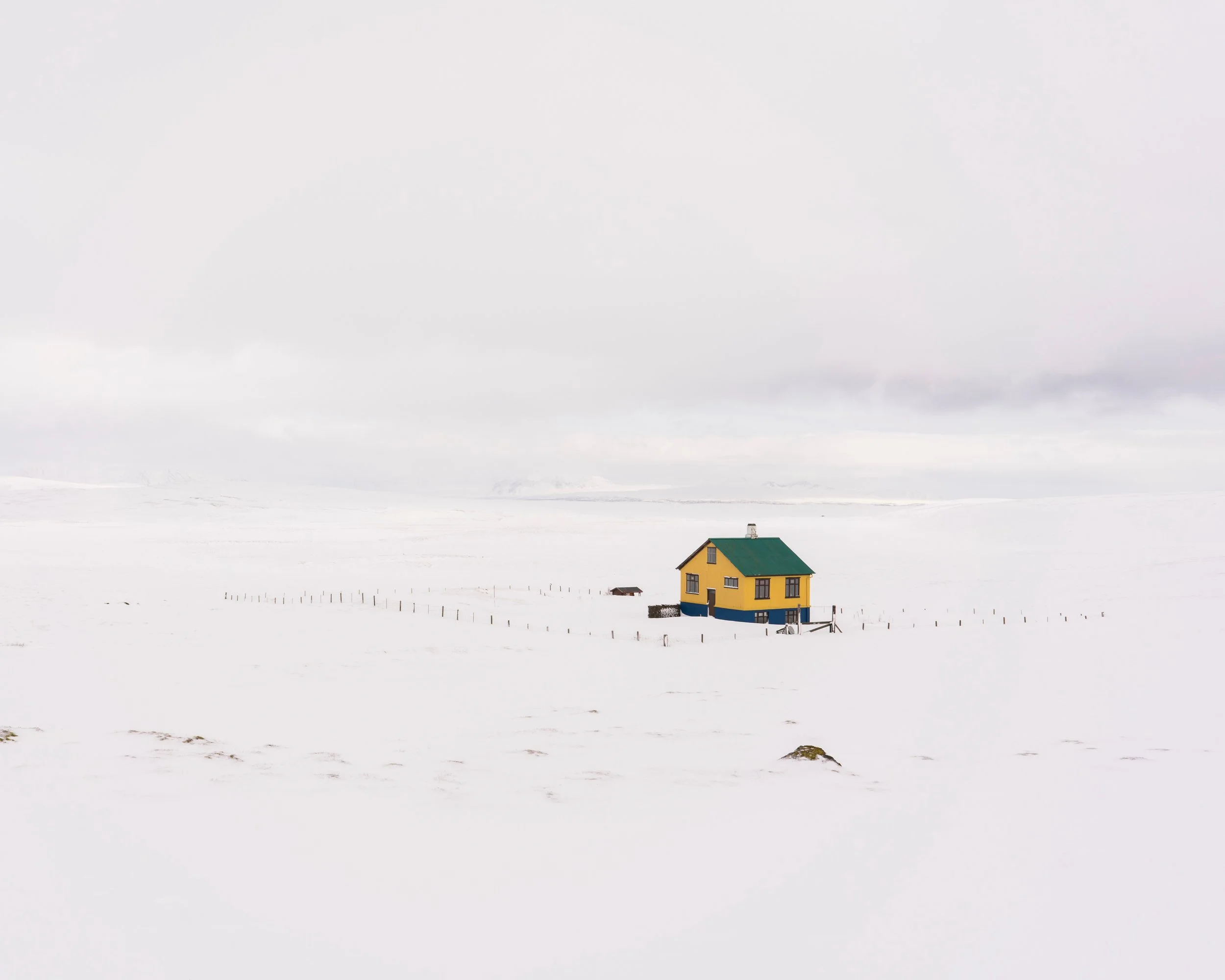 A minimalist fine art photograph capturing a solitary, small yellow house with a bright green roof and blue foundation, surrounded by a simple wooden post fence line in a vast, snow-covered winter landscape in Iceland under an overcast sky.