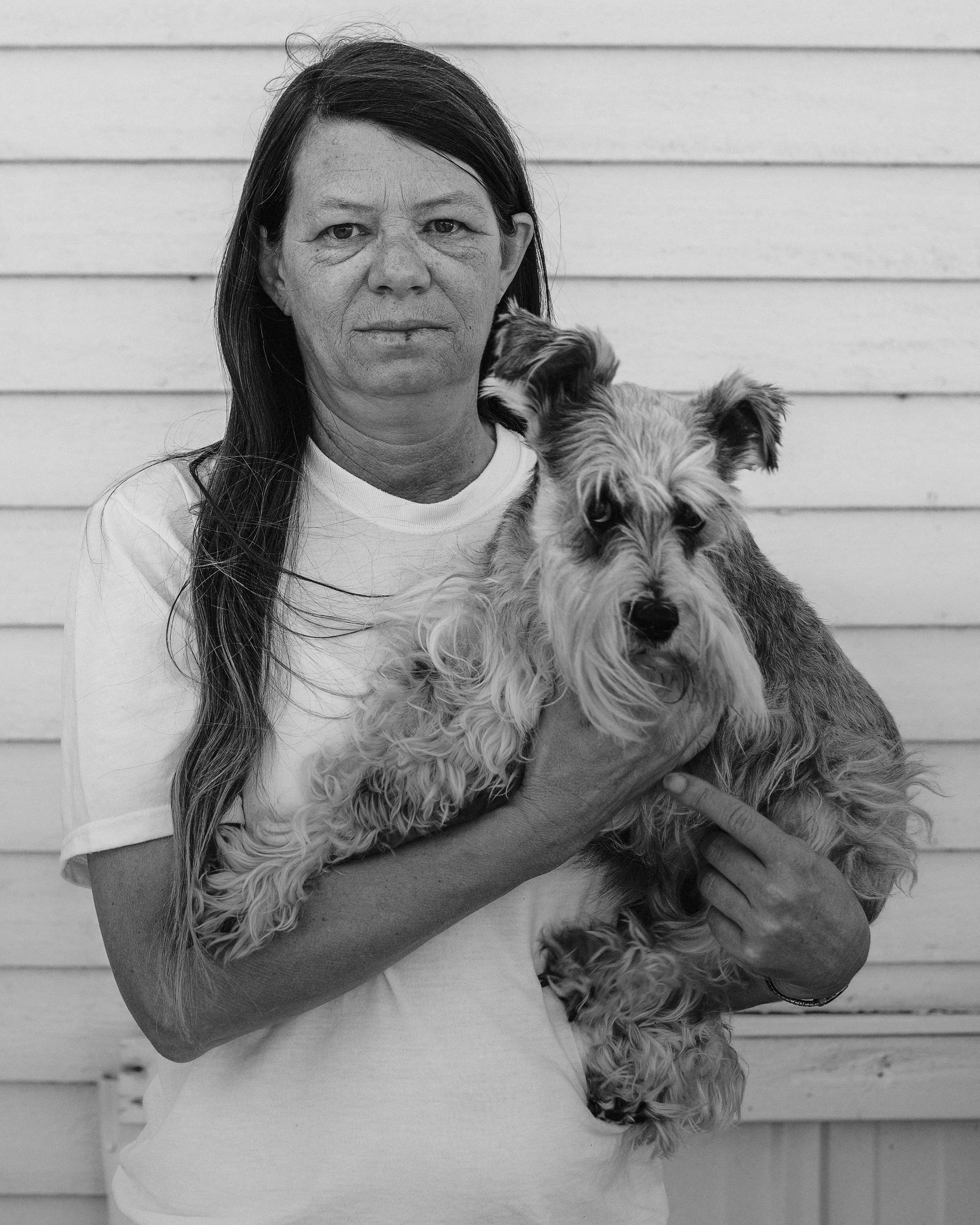 A vertical black and white portrait of a woman lovingly holding a Schnauzer dog in her arms against a light siding background, from One Way Out, Lord by Parker J Pfister.