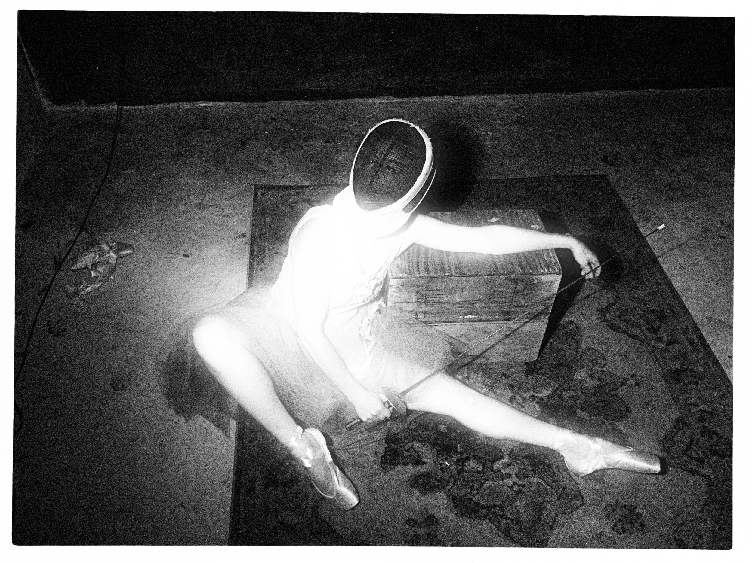 A high-contrast fine art photograph taken from an overhead perspective, showing a ballerina in a tutu and pointe shoes seated on a patterned rug, wearing a dark fencing mask and holding a foil.