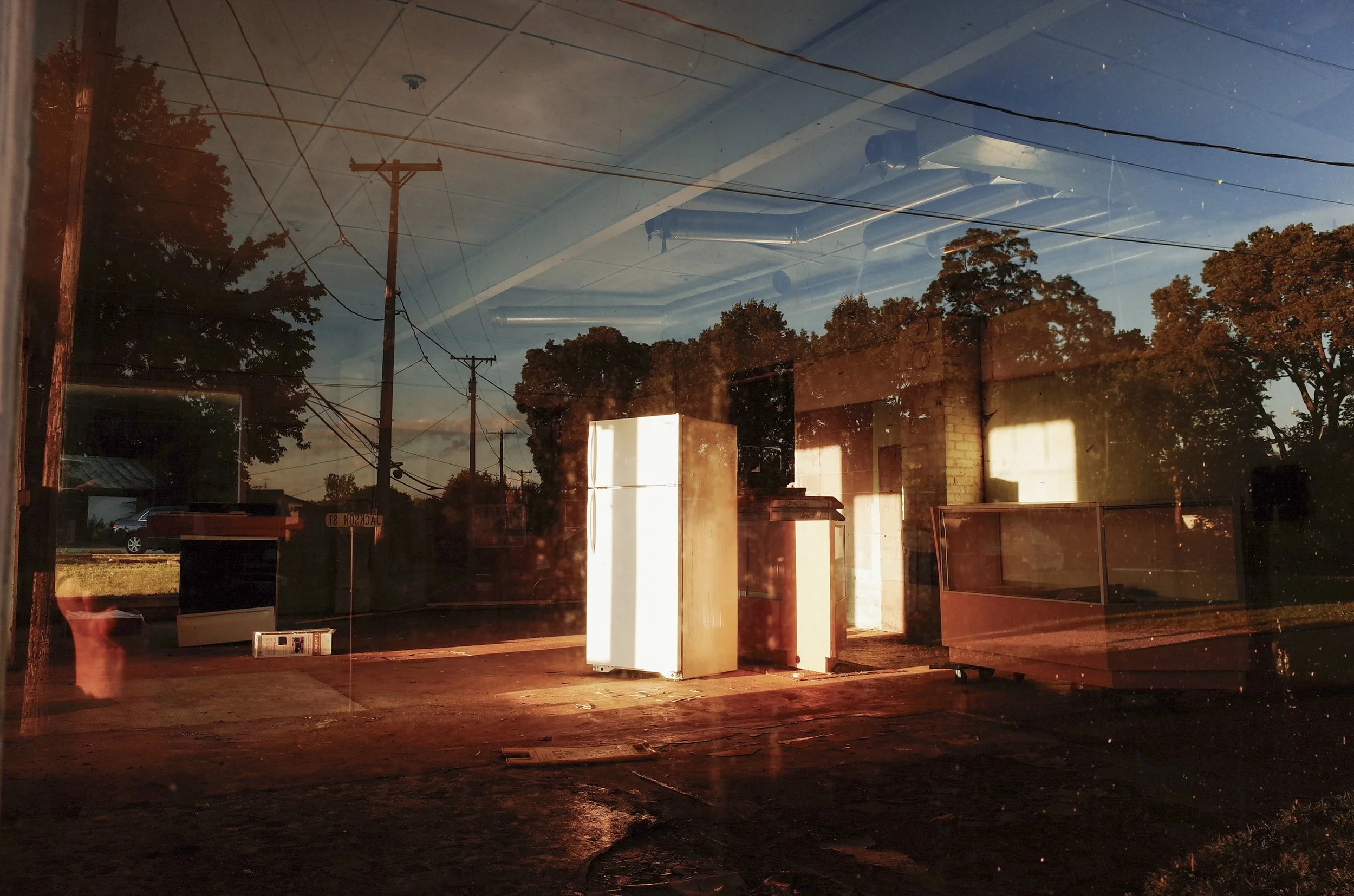 A complex color film photograph using window reflections to layer an old white refrigerator standing in a sunlit lot with power lines and trees, from the One Way Out, Lord series by Parker J Pfister.