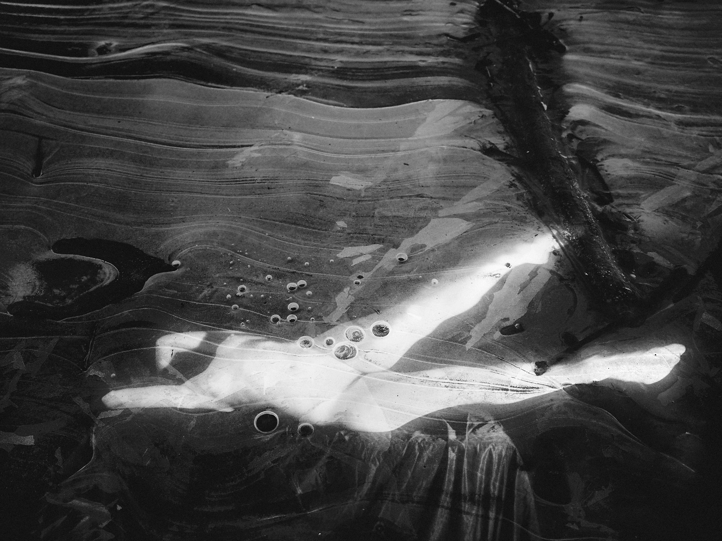 An atmospheric, dark monochrome in-camera double exposure featuring a glowing nude figure seemingly submerged or floating beneath a heavily textured, cracked outdoor ice surface.