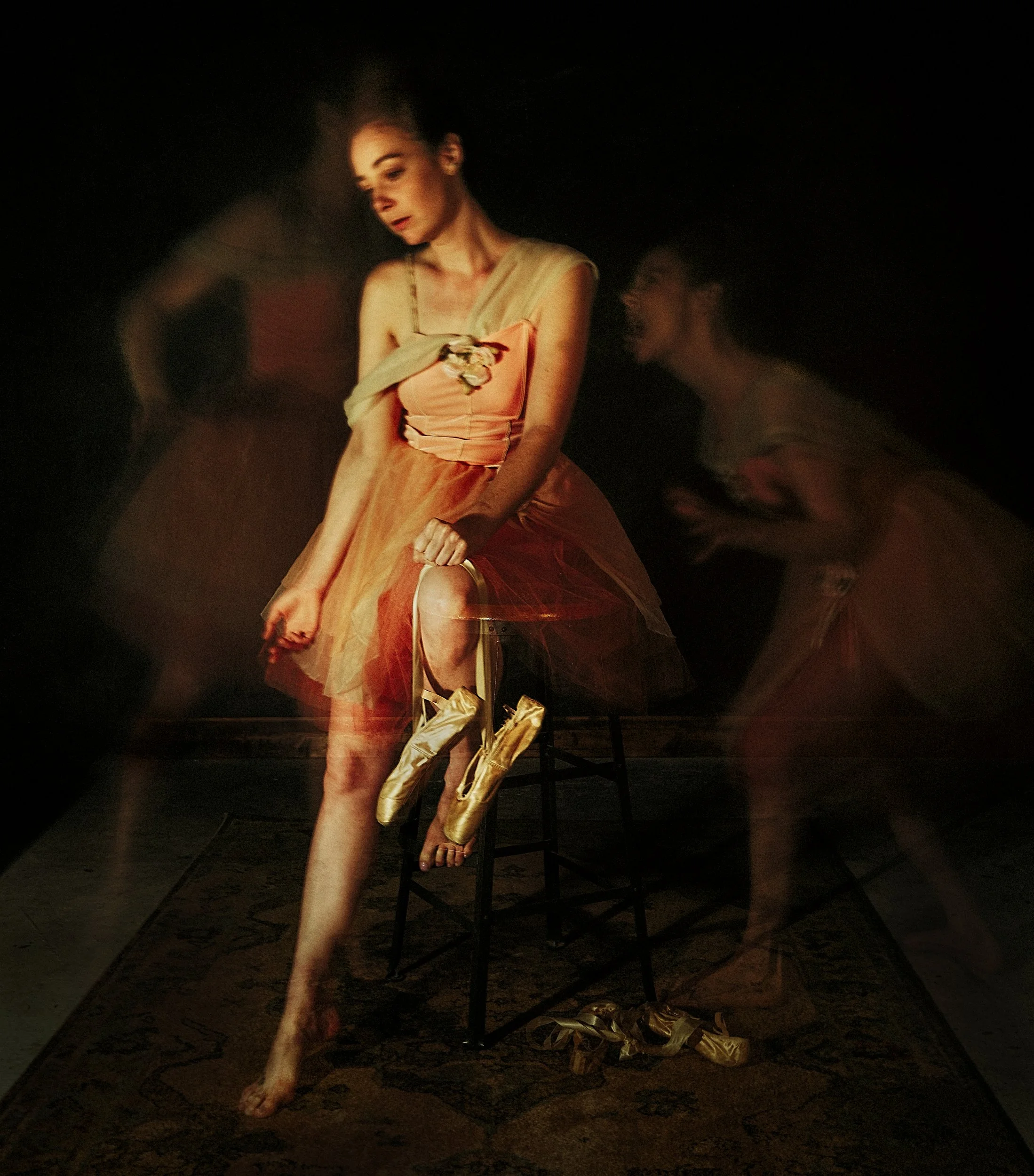A fine art monochrome multiple exposure photograph of a ballerina sitting on a stool holding pointe shoes, surrounded by ghostly, layered motion frames that evoke a sense of struggle.