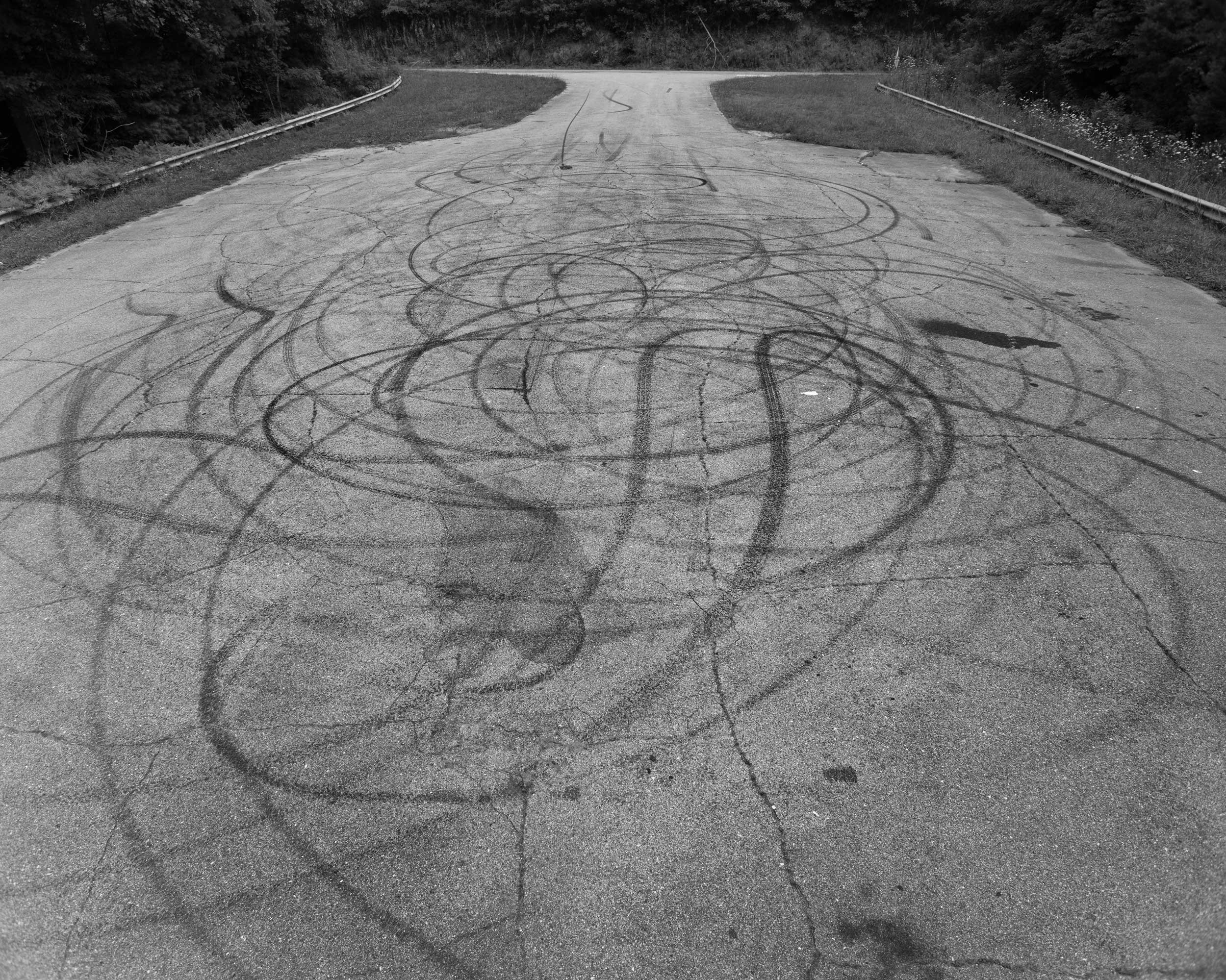 A high-angle black and white documentary photograph showing intricate, overlapping circular tire burnout marks layered across the asphalt of a quiet, curving mountain road in Western North Carolina.