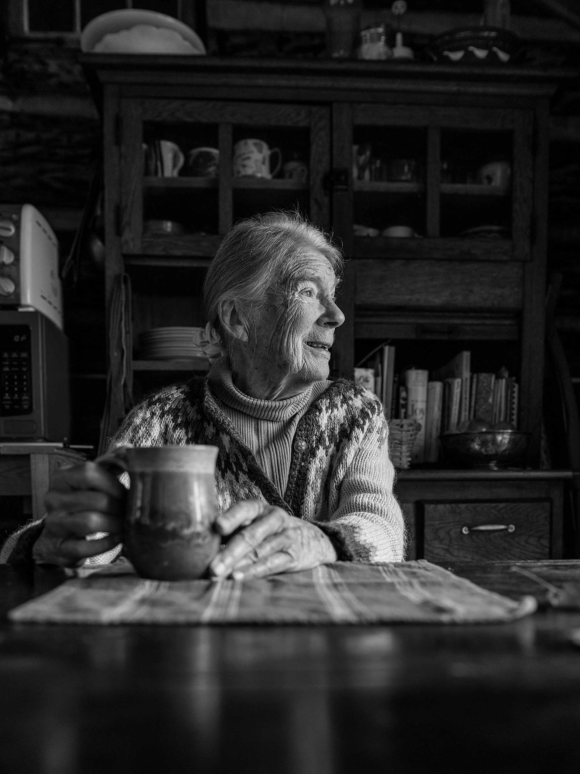 Black and white documentary portrait of an elderly Appalachian woman sitting at a table, holding a ceramic mug and looking out a window, with a rustic wooden hutch filled with books and dishes in the background of her Western North Carolina home.