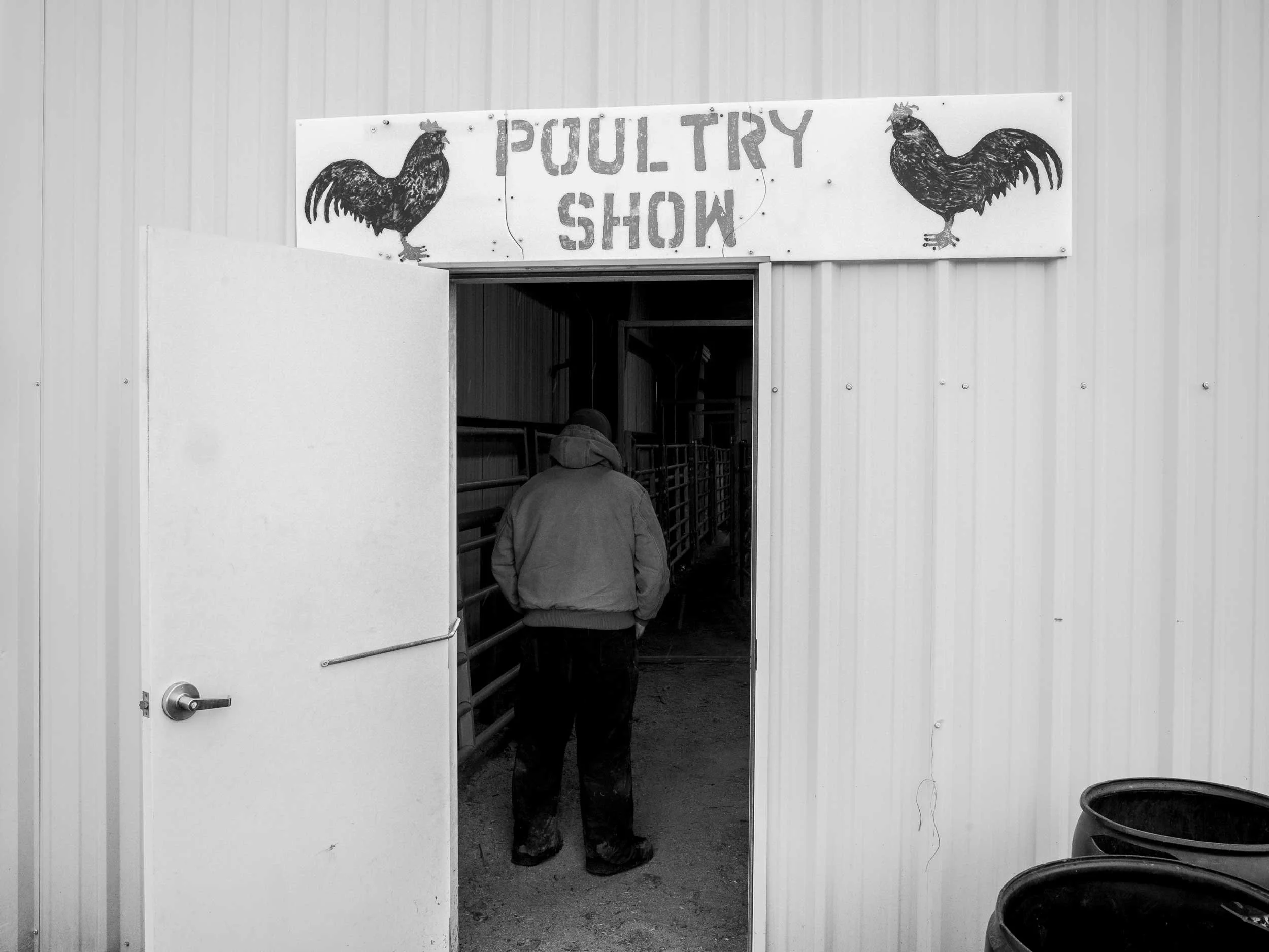 A high-contrast black and white documentary photograph showing the back of a person walking into a building doorway under a hand-painted sign that reads "POULTRY SHOW" with drawings of roosters in Western North Carolina.