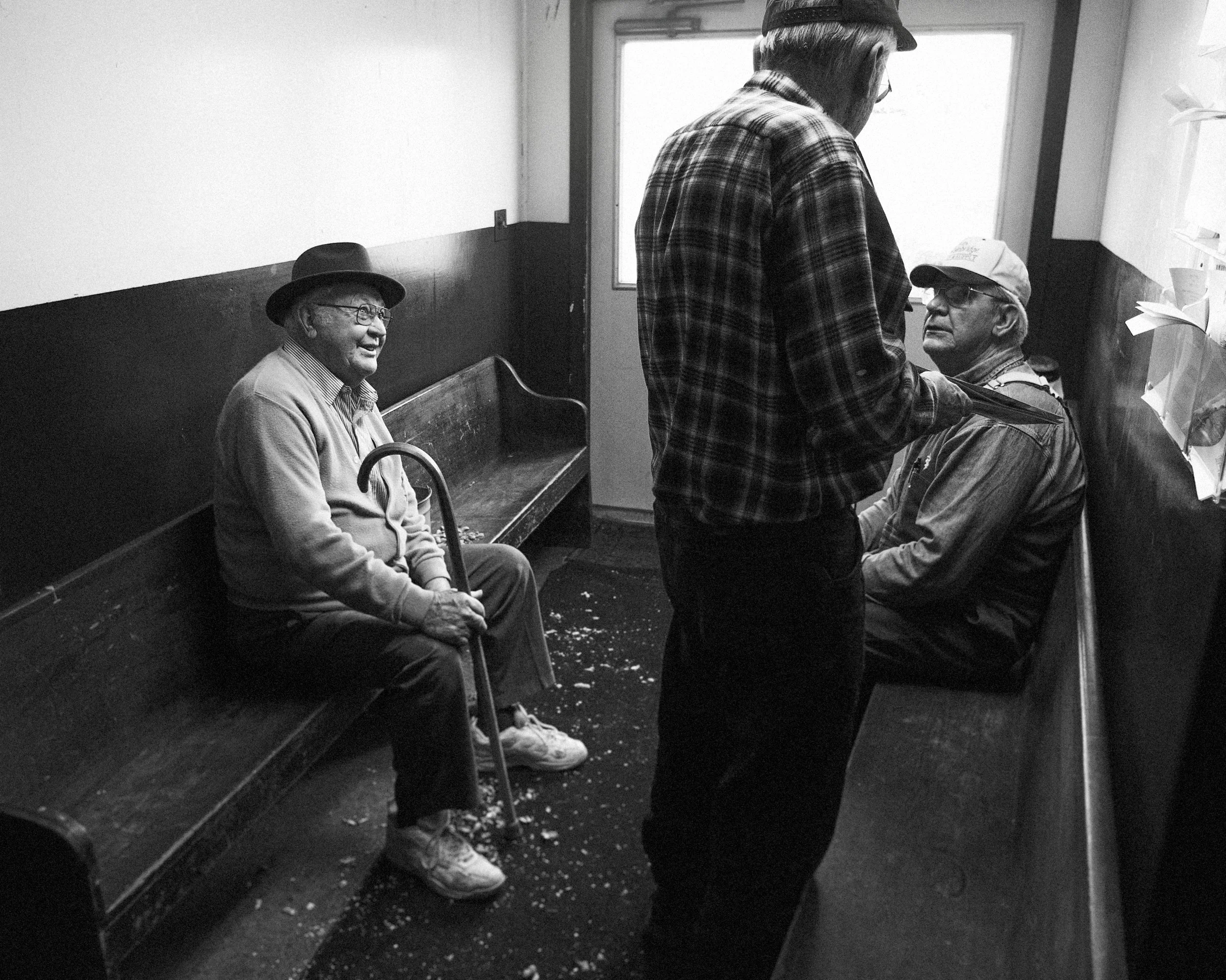 A black and white environmental photograph of older men talking on wooden benches inside the Union Stockyards in Hillsboro, Ohio, with discarded peanut shells covering the floor, from One Way Out, Lord by Parker J Pfister.