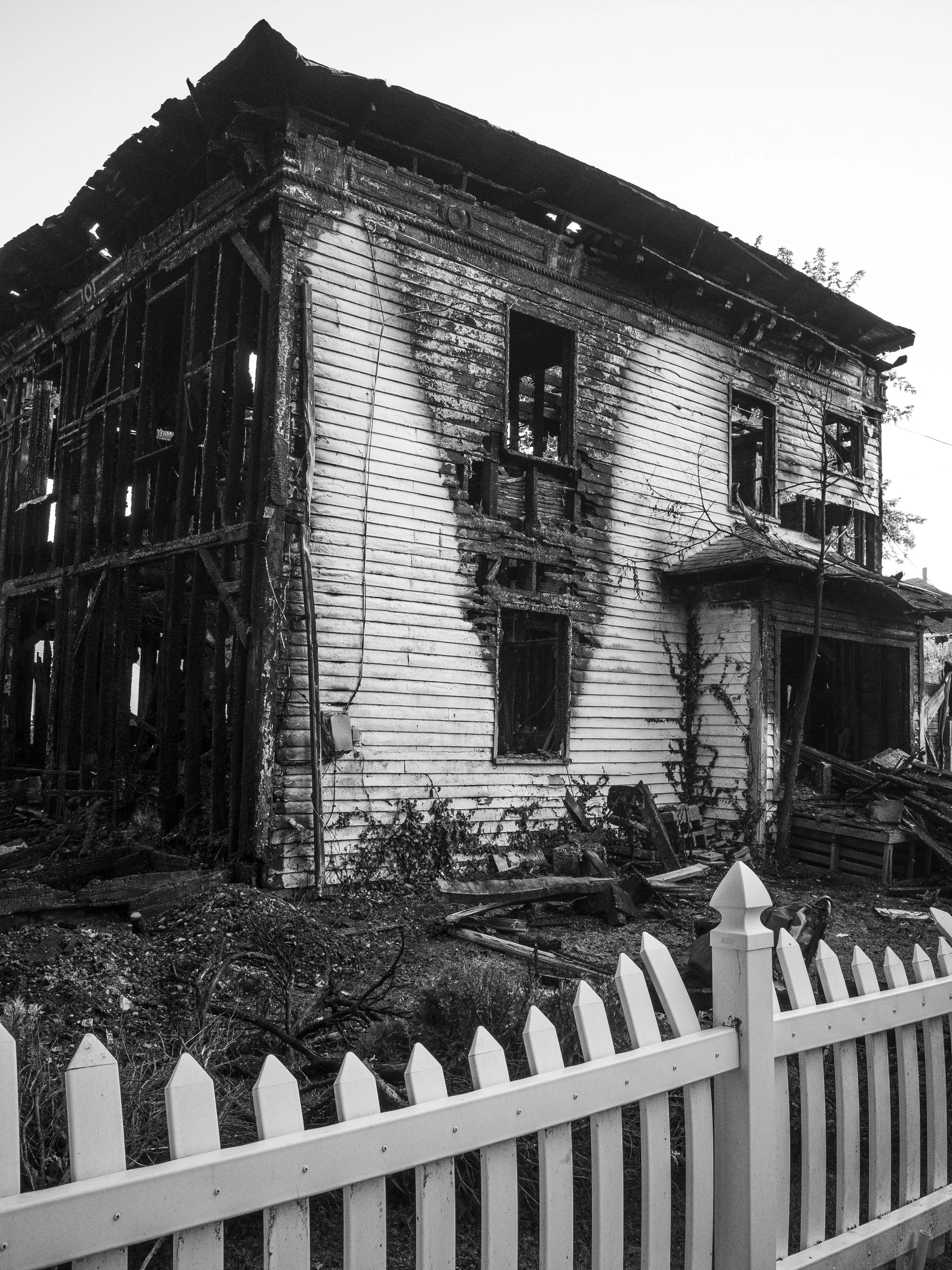 A powerful black and white documentary photograph of a two-story residential house destroyed by fire, with a white vinyl picket fence melted and warped from the heat in the foreground, from One Way Out, Lord by Parker J Pfister.