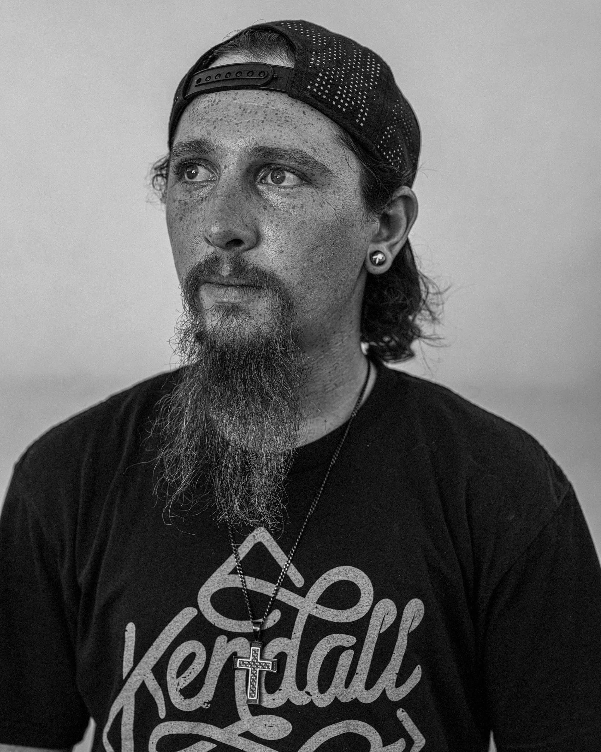 A crisp, detailed digital black and white portrait of a man with a long goatee beard and freckles, wearing a backward baseball cap and a "Kendall" t-shirt, captured at the Madison County rodeo in North Carolina.